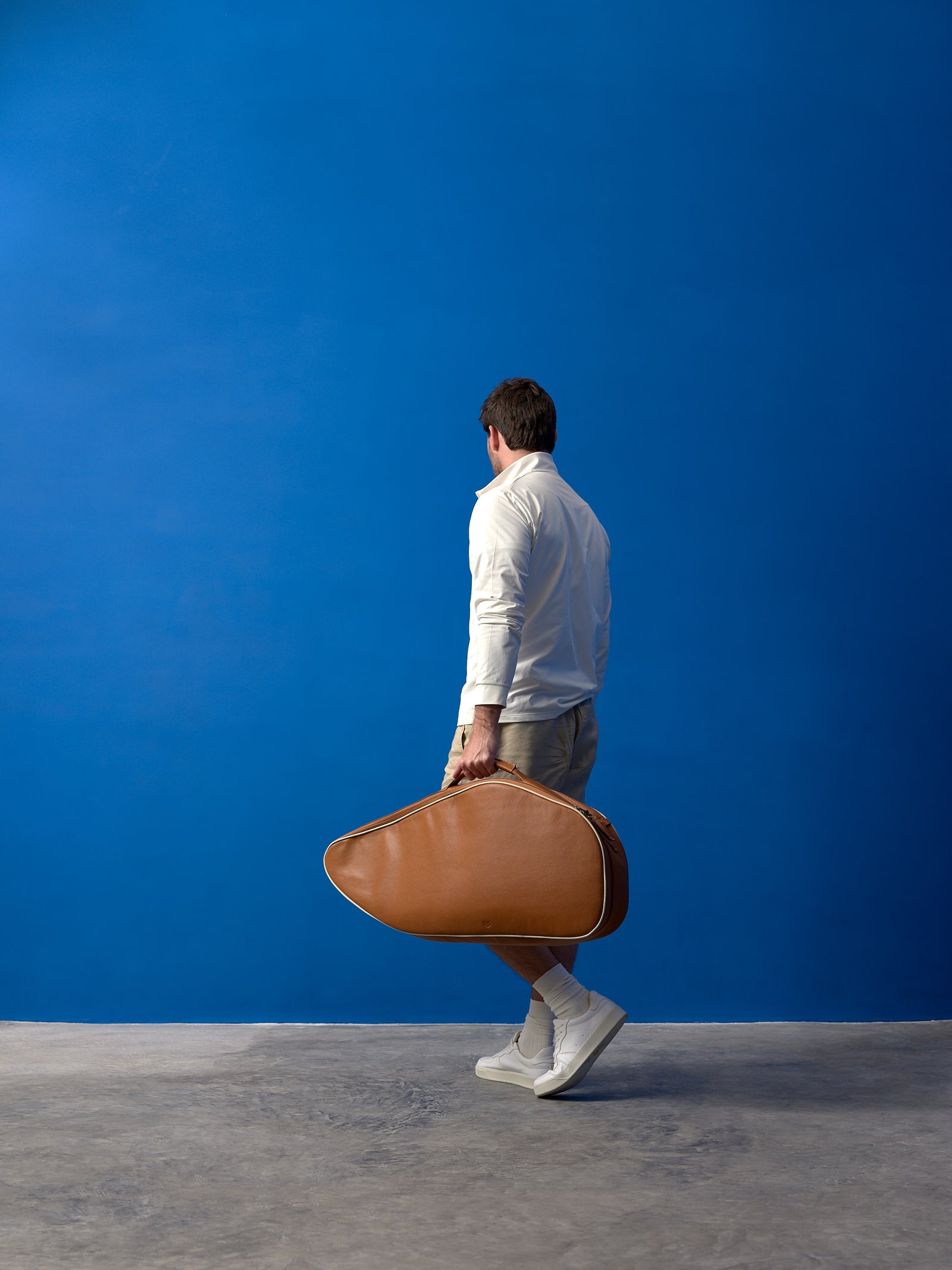 Back view of a man walking in a blue studio, carrying a large tan leather tennis bag by the side handle.