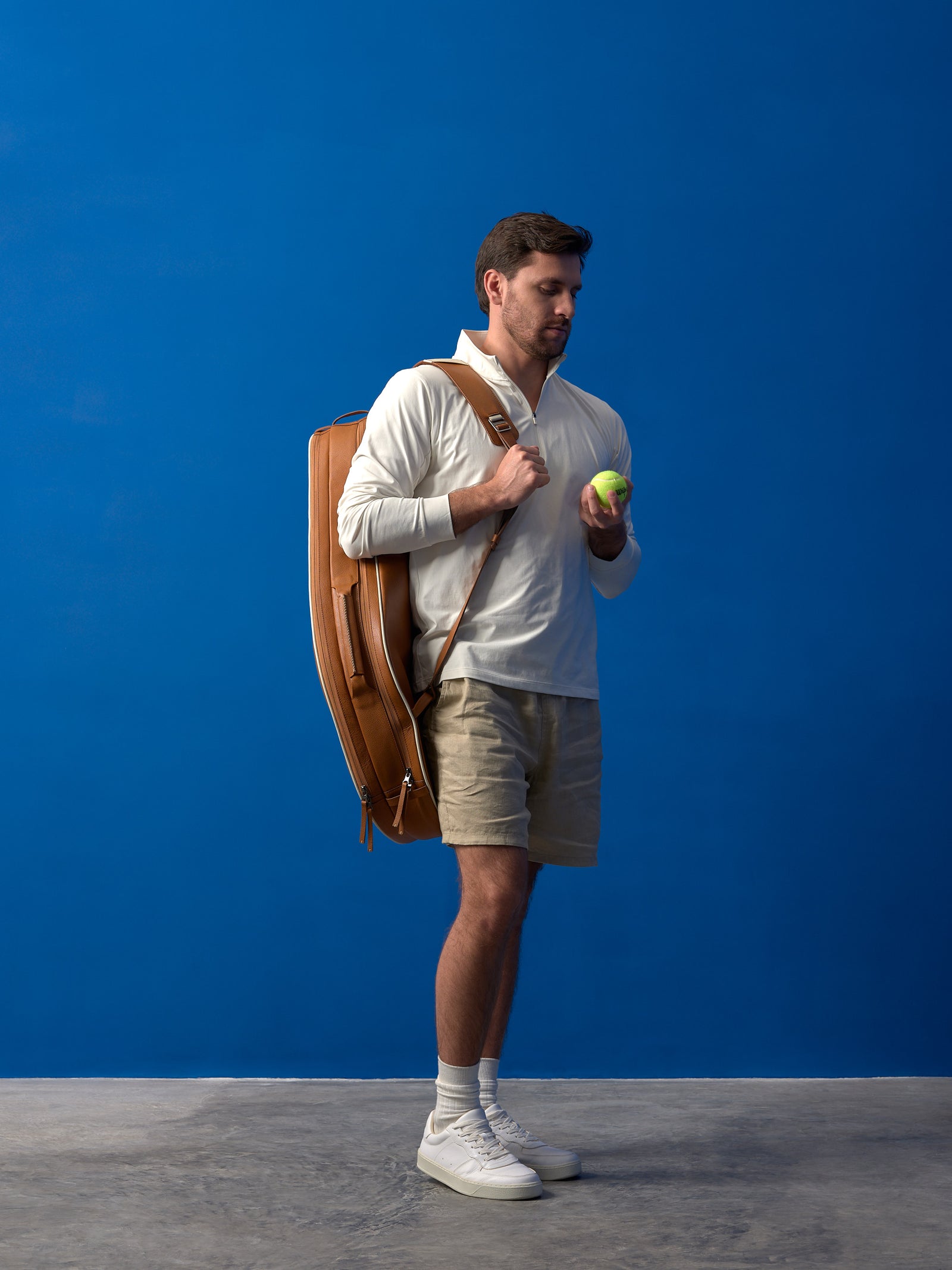 Full-body view of a male player wearing a cream pullover and holding a tennis ball, with a tan leather bag over his shoulder.