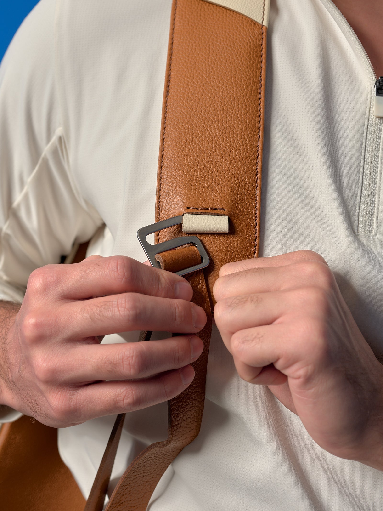 Close-up of hands adjusting the padded tan full-grain leather strap and metal buckle of a Capra Leather tennis bag.