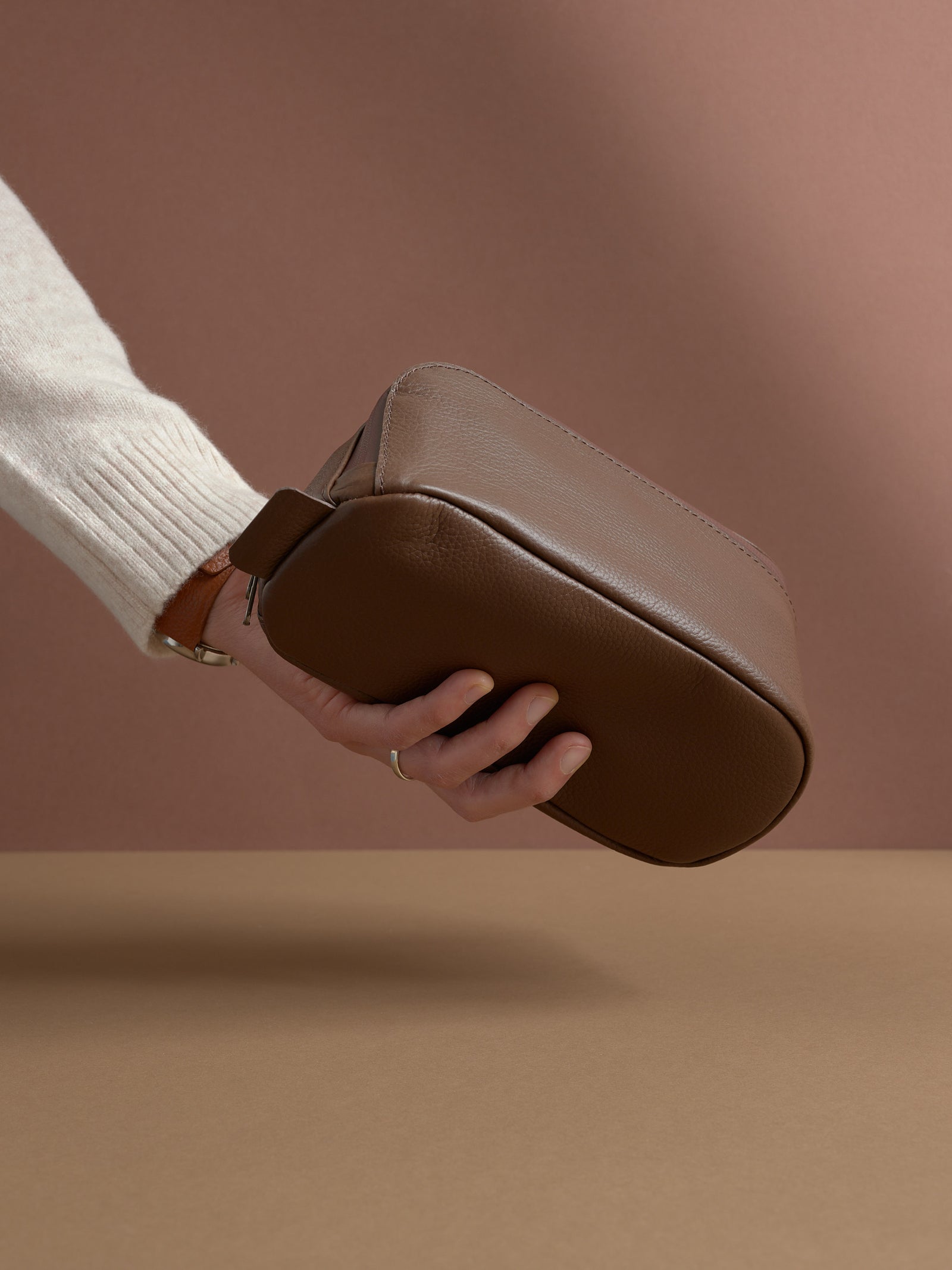 A hand holding the closed brown Capra Leather Toiletry Bag by the side profile.