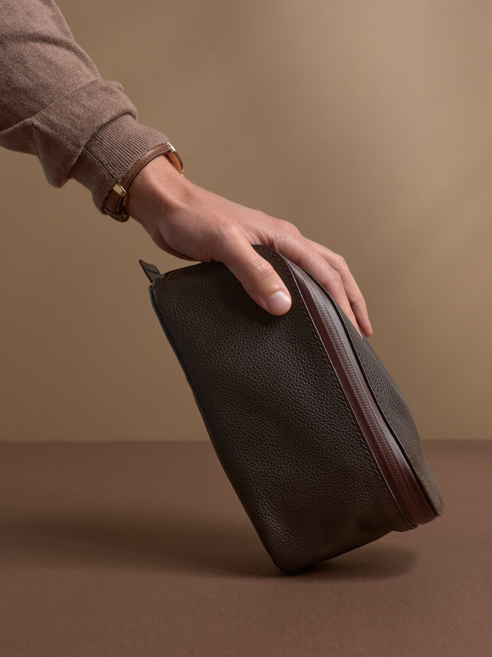 A hand resting on the standing dark brown Capra Leather Toiletry Bag.