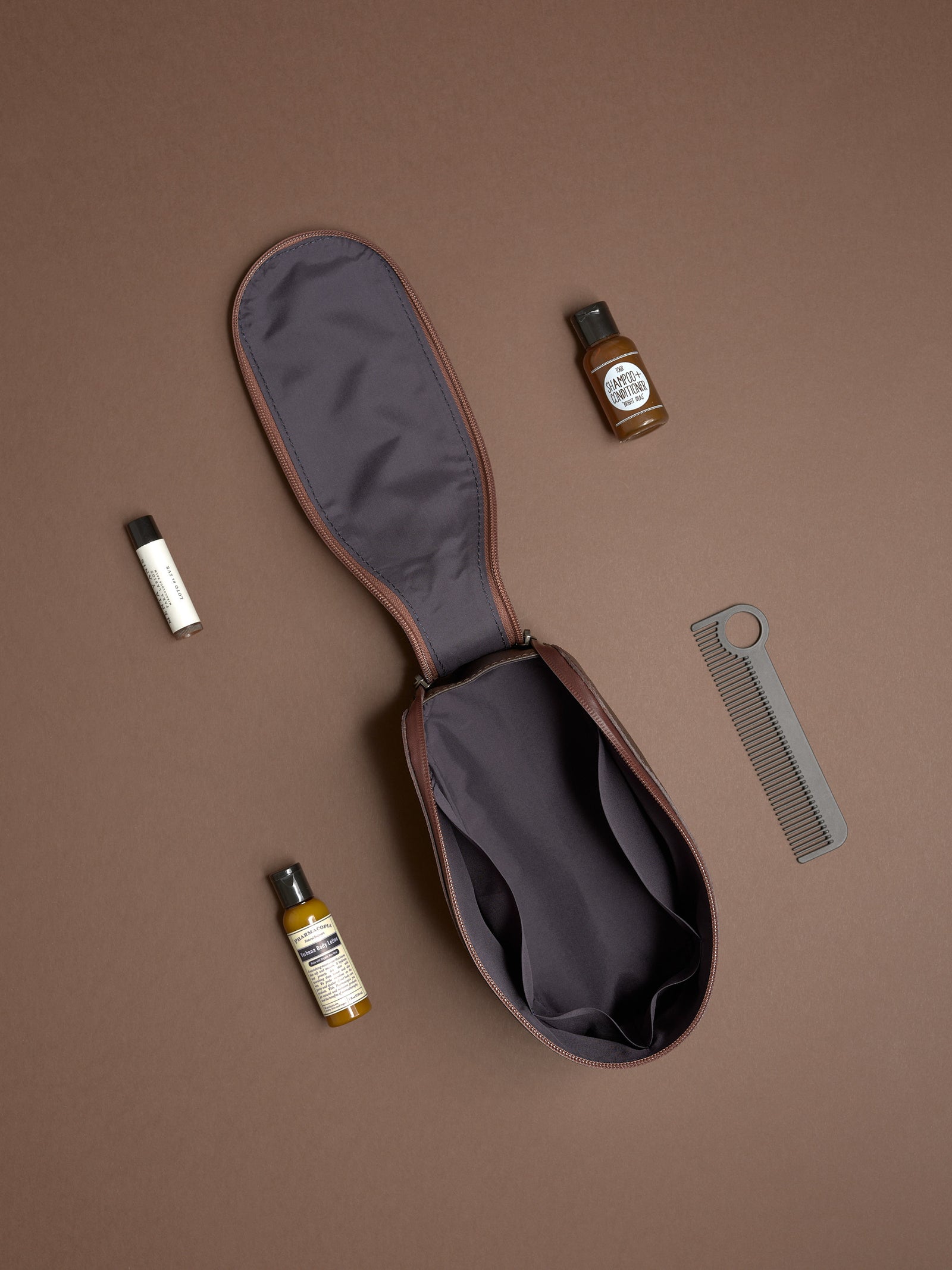 Open dark brown Capra Leather Toiletry Bag laid flat with various travel grooming products.