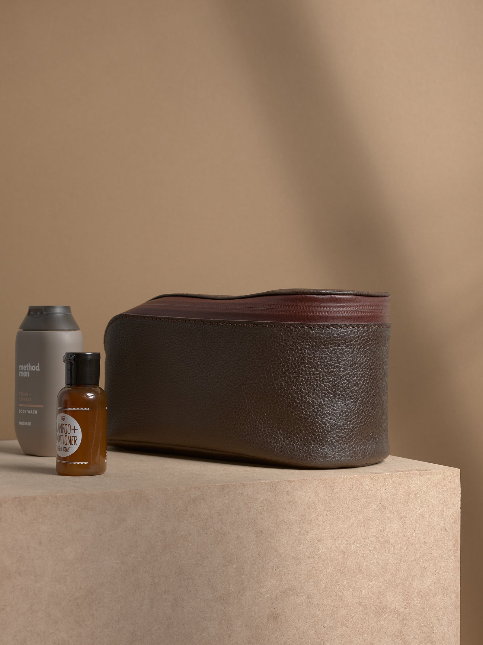 Closed dark brown Capra Leather Toiletry Bag next to small bottles of shower products.