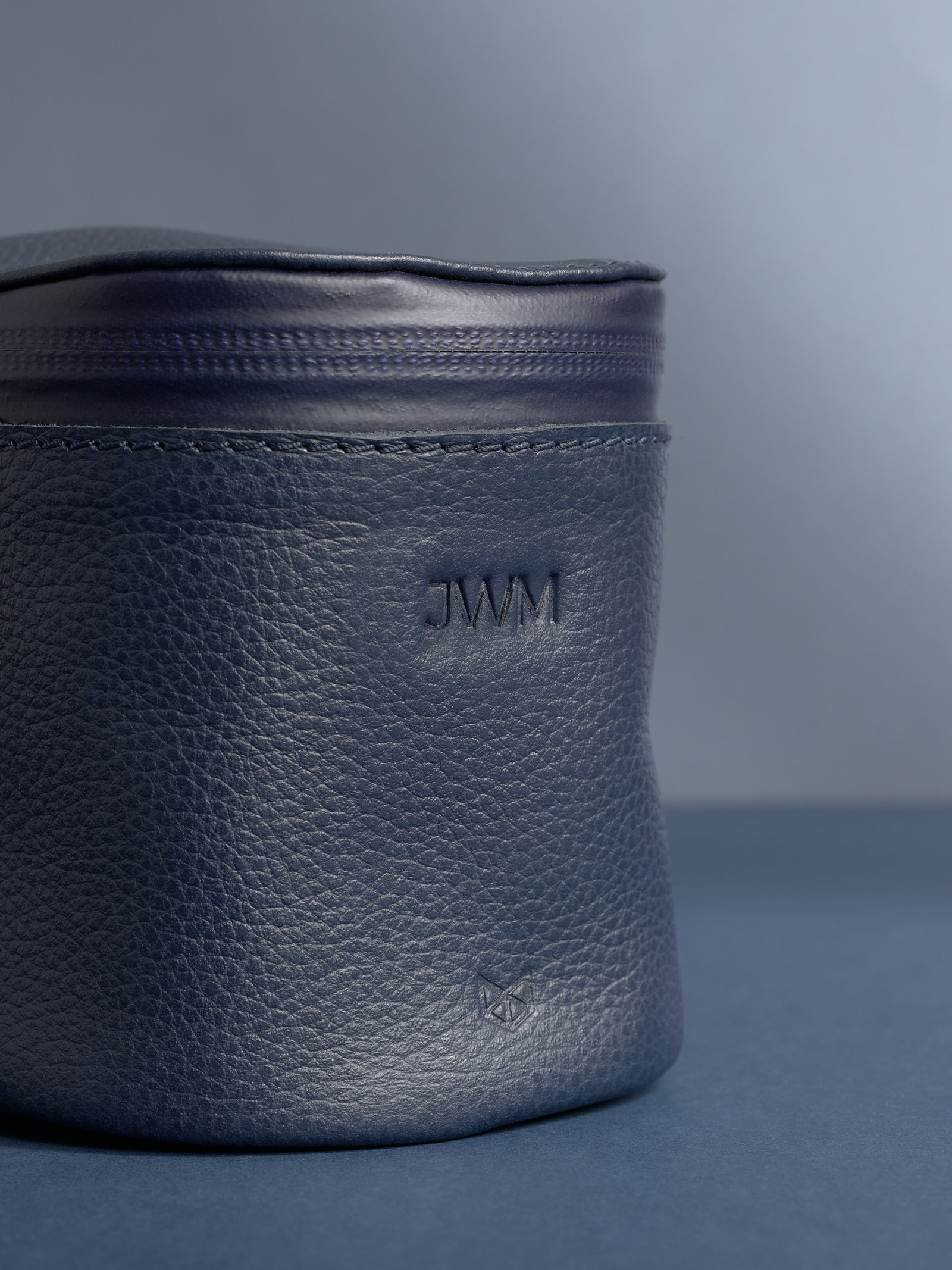Close-up of a personalized navy blue Capra leather cylindrical toiletry bag. The leather has a pebbled texture, and the initials "JWM" are debossed near the top.