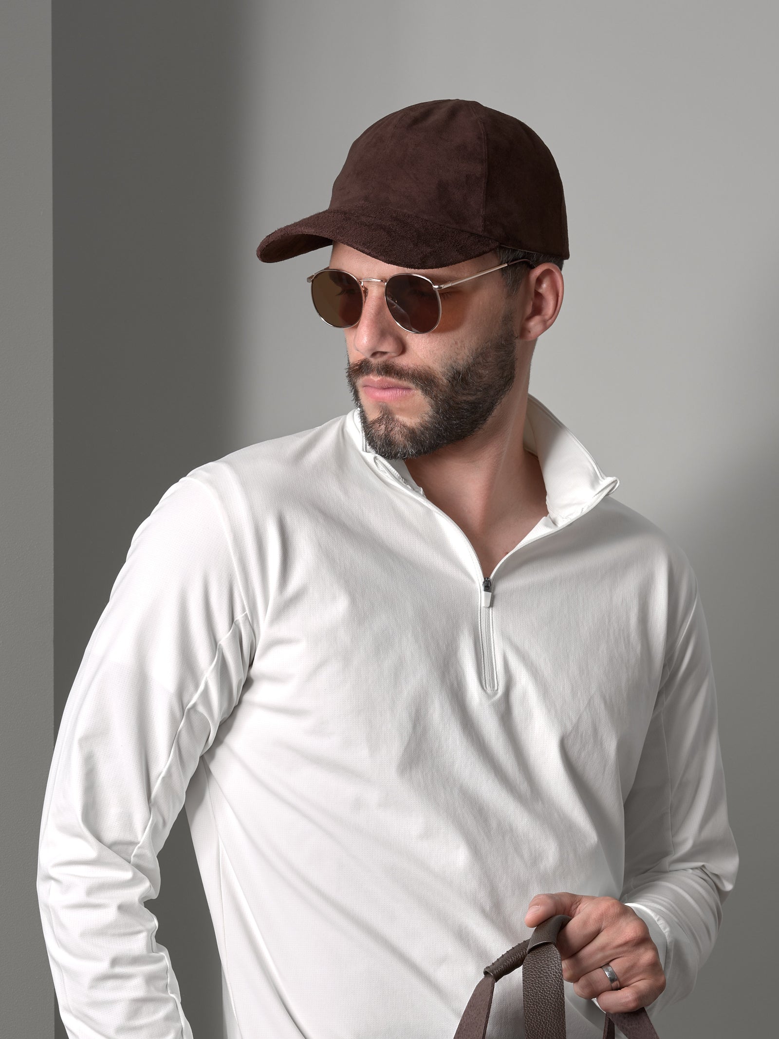 A stylish man wearing a brown leather suede cap, gold aviator sunglasses, and a white quarter-zip shirt.