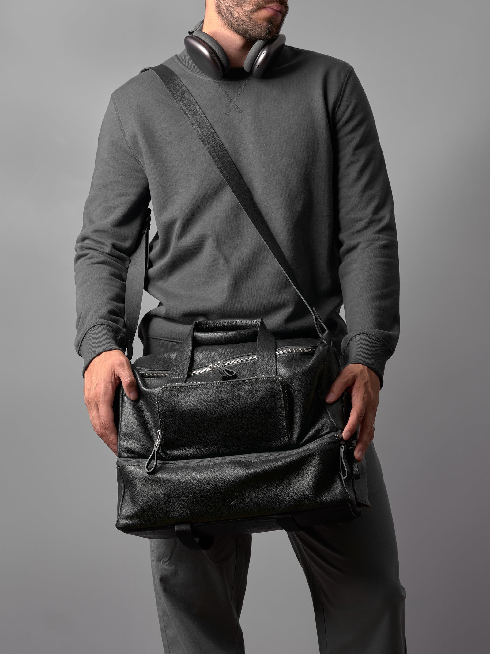 A man wearing headphones carries a black leather Capra duffel, showcasing its adjustable shoulder strap.