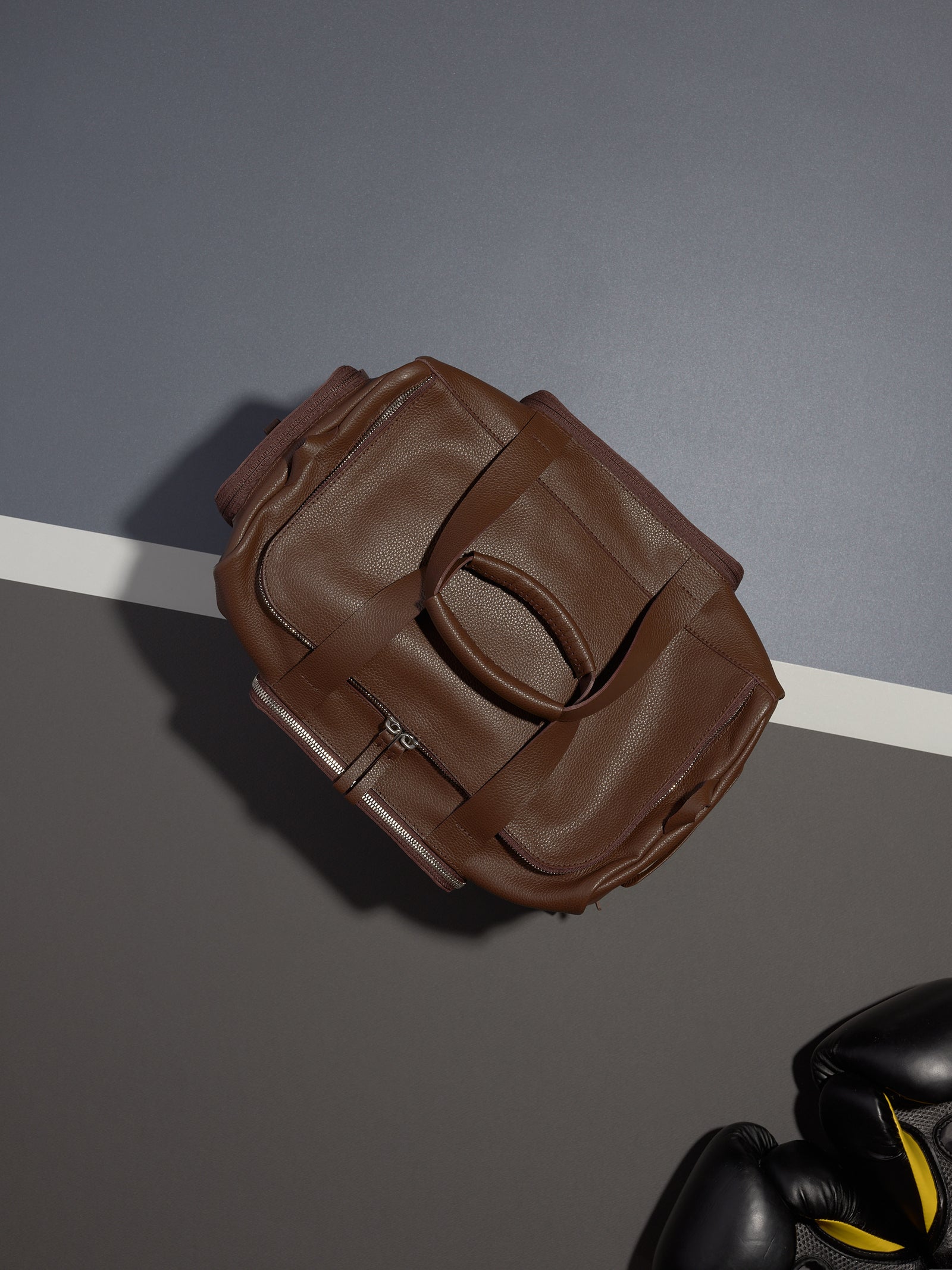 A top-down shot of the brown leather Ventra Holdall next to a pair of boxing gloves on a color-blocked surface.