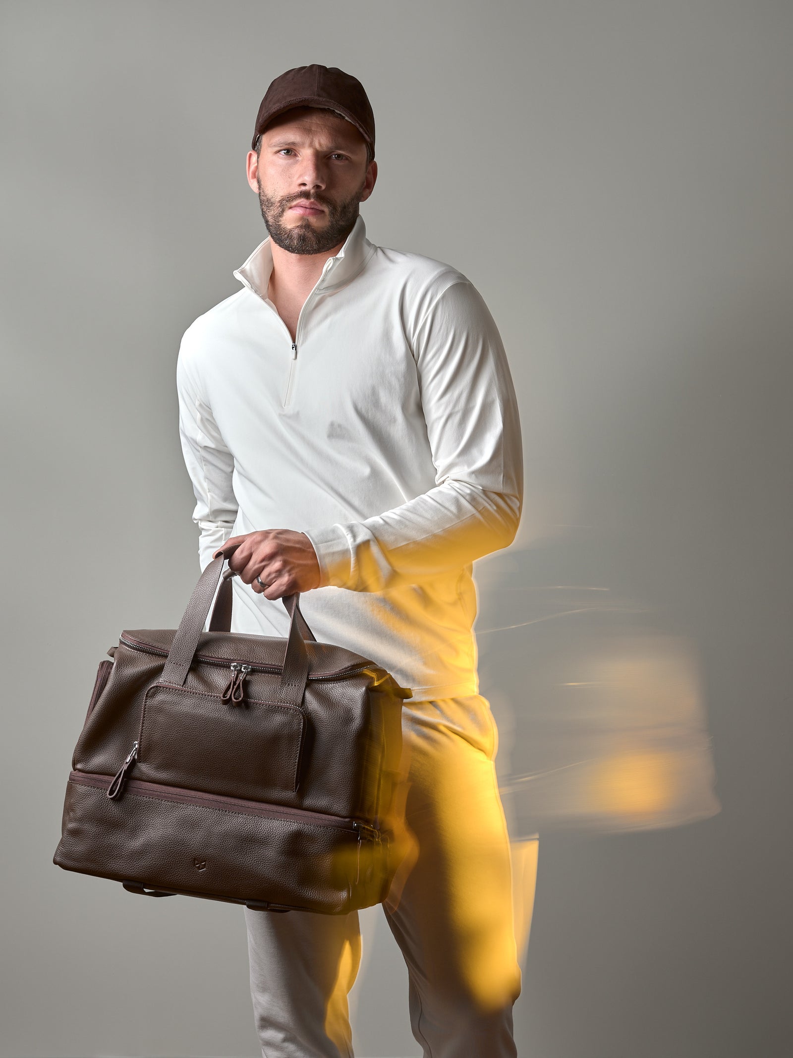 A man in a white long-sleeve shirt and cap holding a dark brown Capra Leather Ventra Holdall duffel bag.