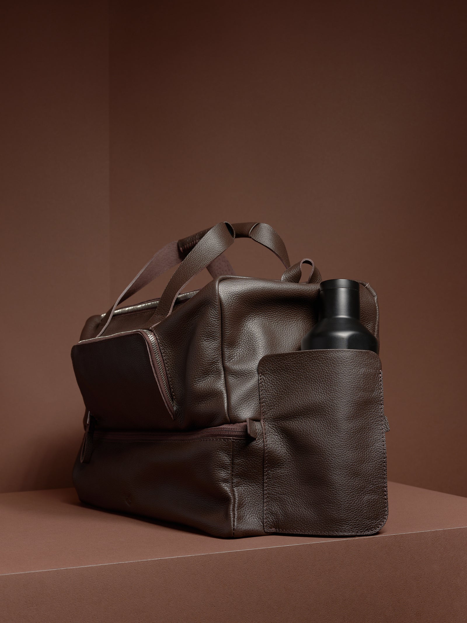 A dark brown leather Ventra Holdall bag on a brown platform with a black water bottle in the side pocket.