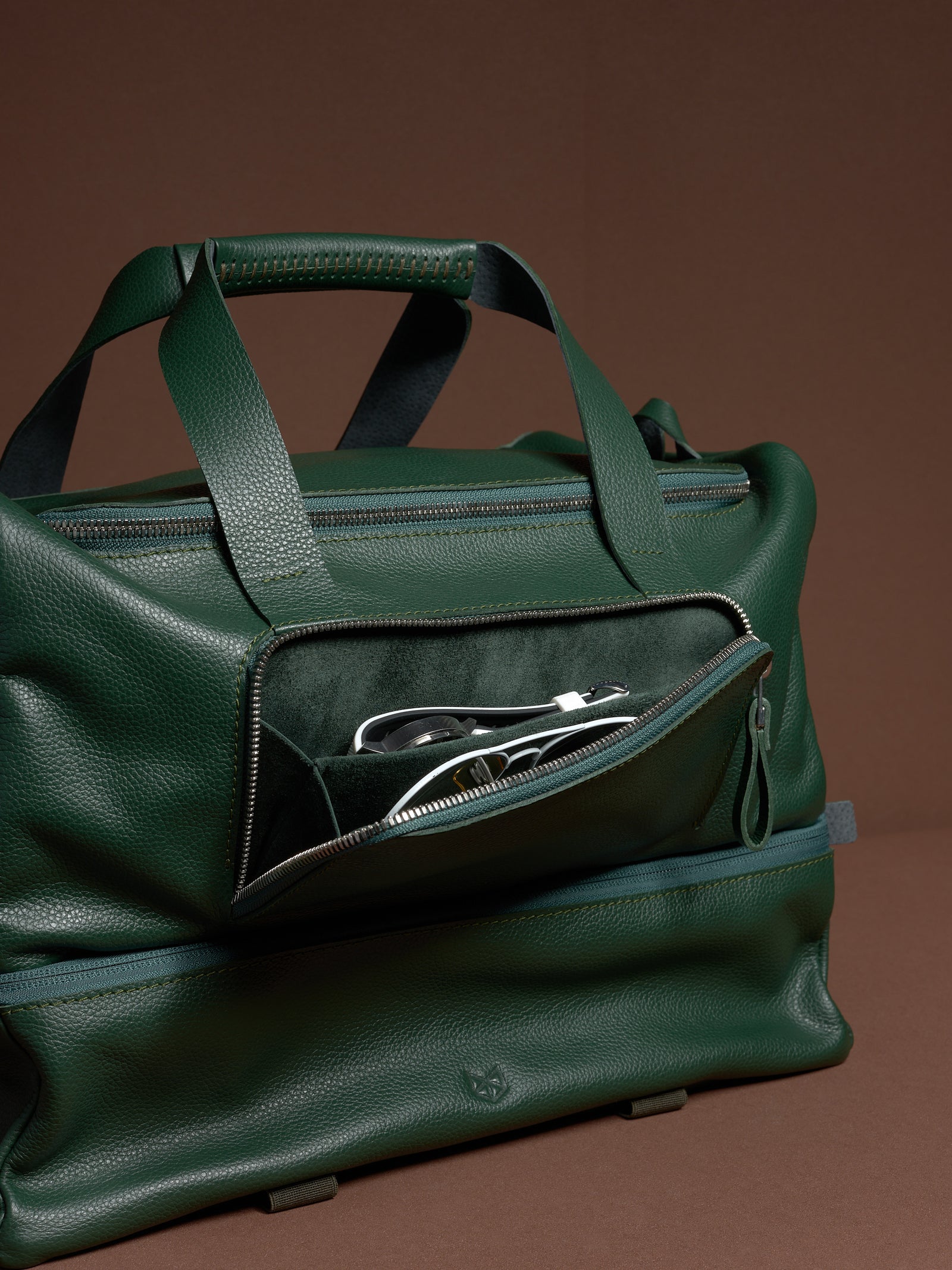 The easy-access front zippered pocket of the green leather Ventra Holdall, open to show wristwatch and glasses inside.