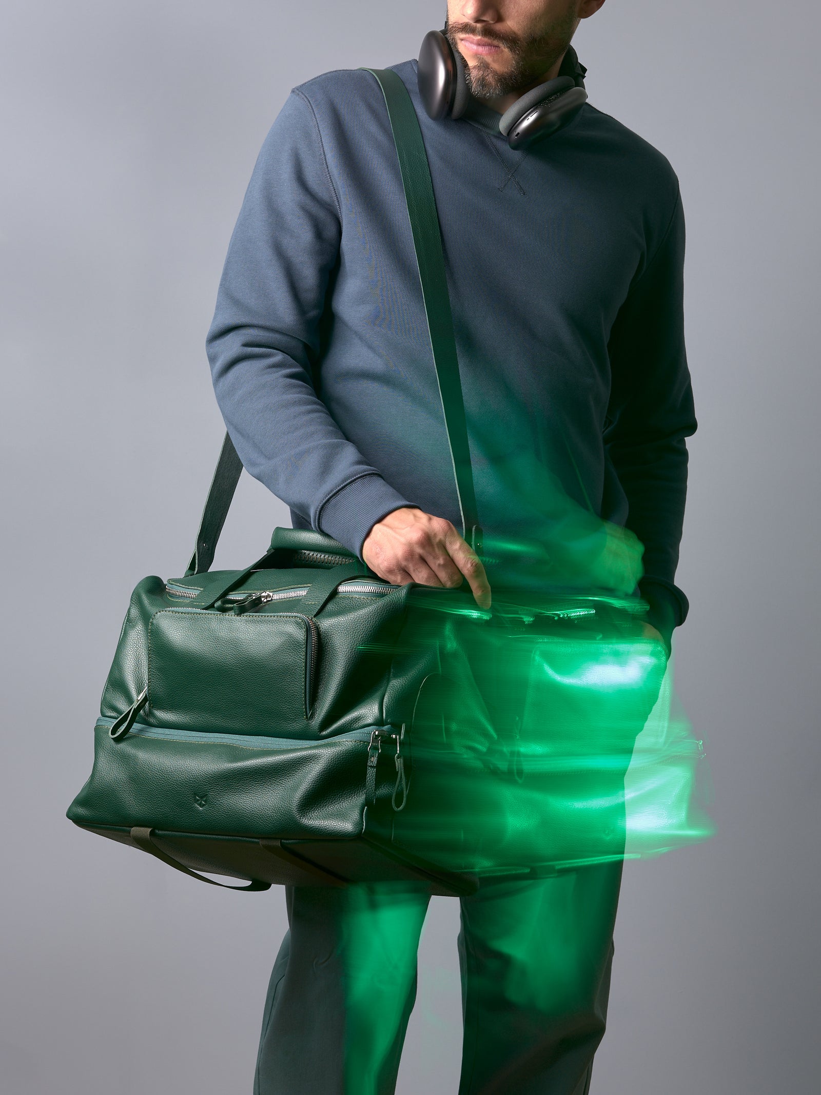 Man wearing headphones and a blue sweatshirt showcasing a green leather duffle bag with a shoulder strap.