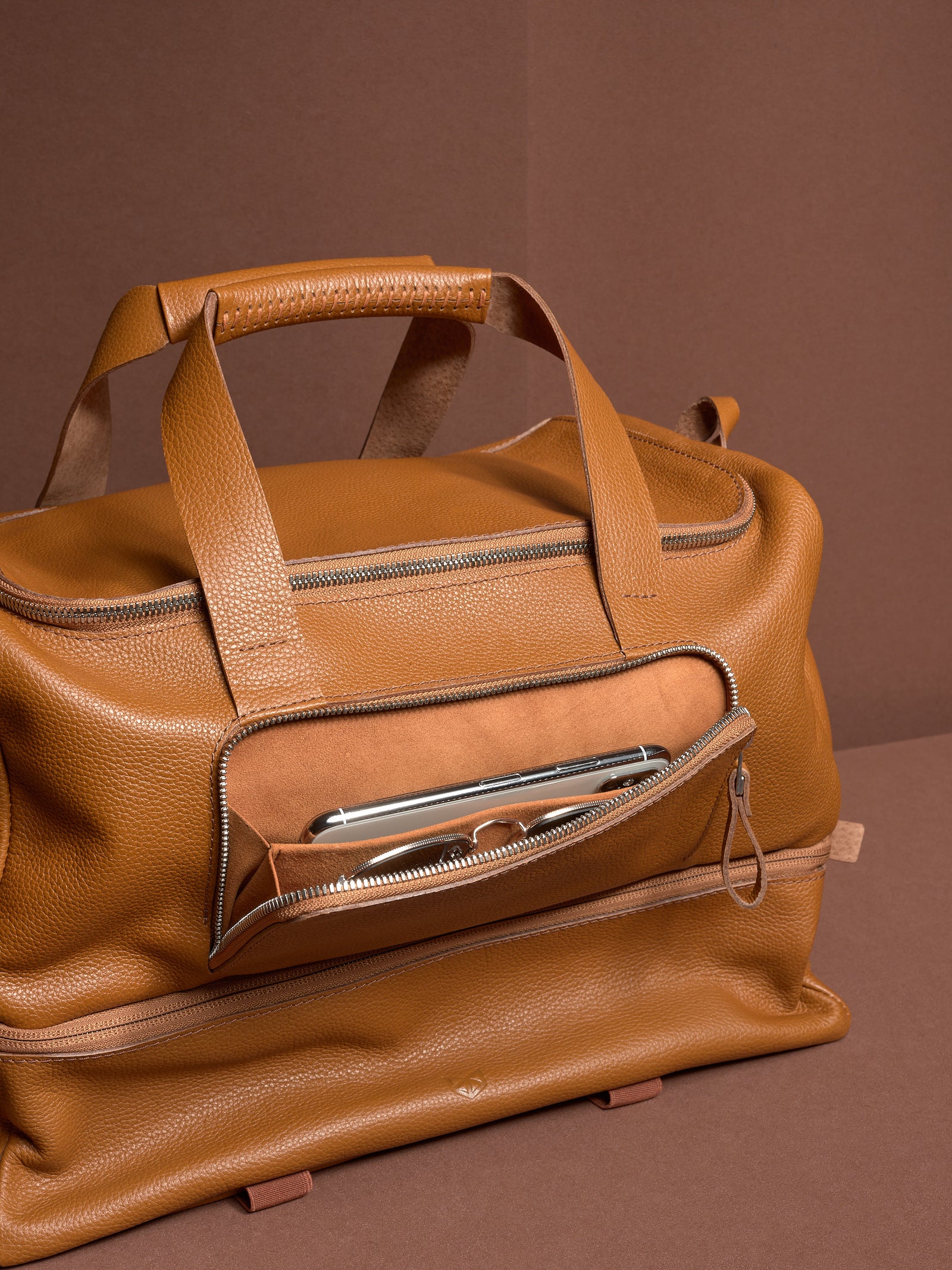 The tan leather Ventra Holdall's front zippered pocket is open, revealing a smartphone and glasses stored for easy access.