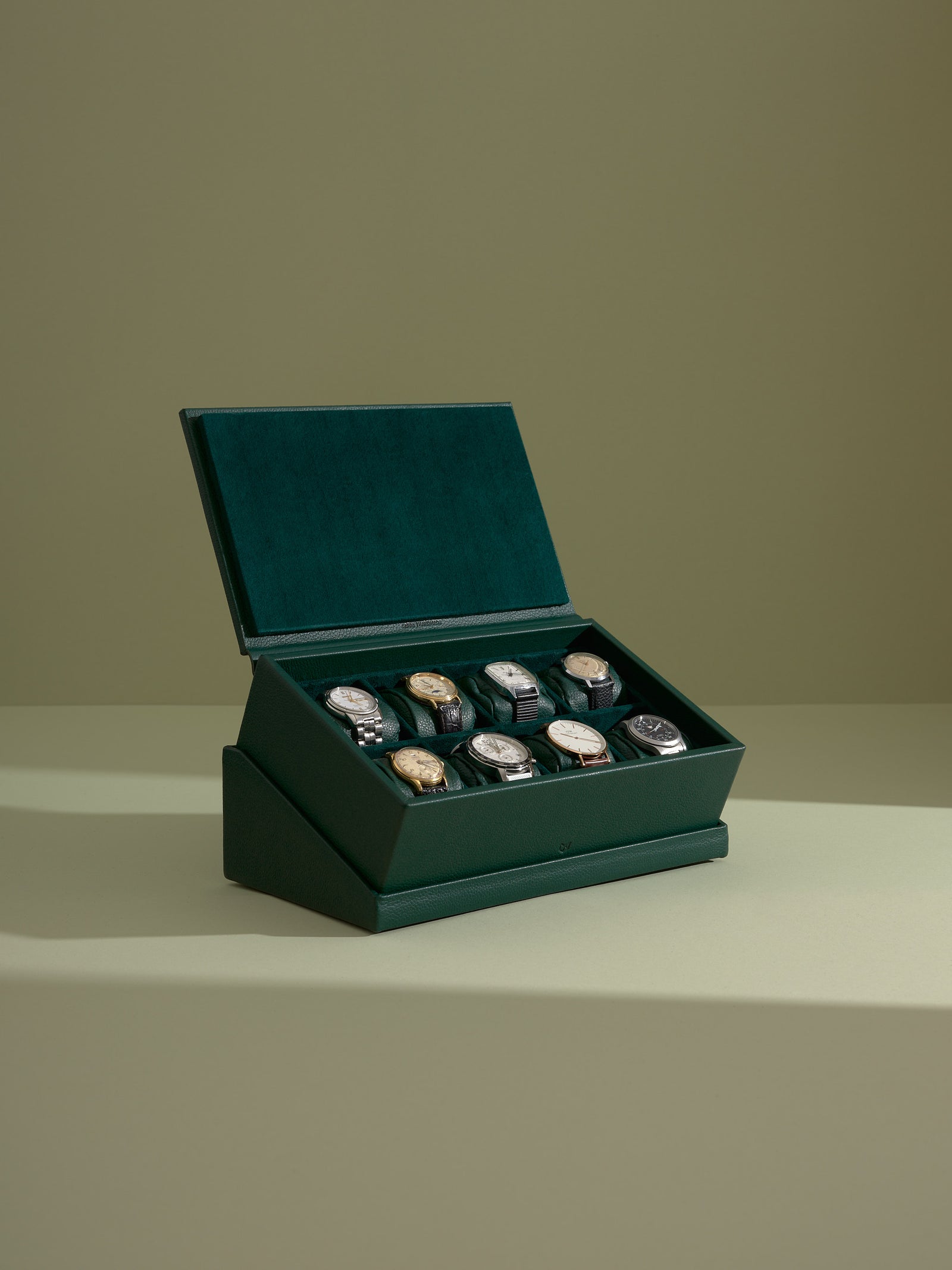 Open green leather watch box presenting eight vintage wristwatches on matching green suede watch pillows.
