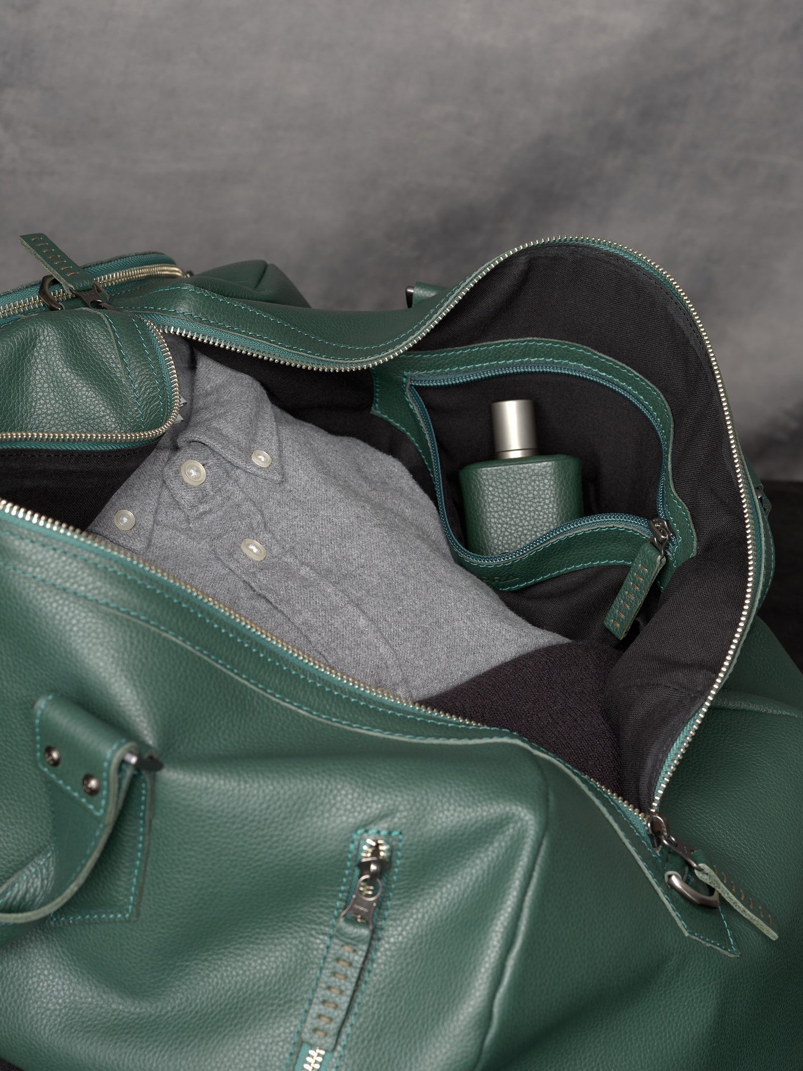 Interior view of the green leather duffle bag packed with a grey shirt and a leather-wrapped fragrance bottle.