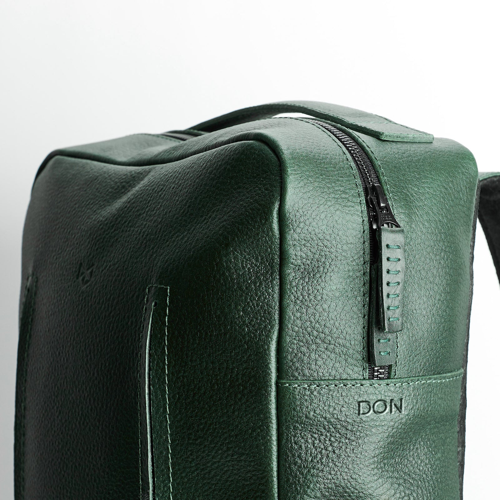 A three-quarter view close-up of the rectangular green leather bag, highlighting the top zipper and the embossed word "DON" near the bottom front panel.