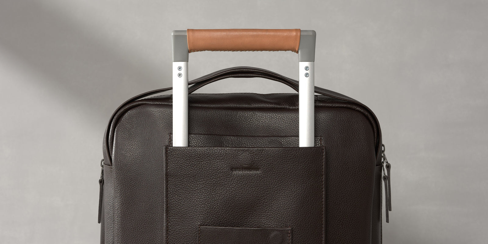 Close-up of a dark brown leather travel bag slid over a silver suitcase trolley handle with a tan leather grip.