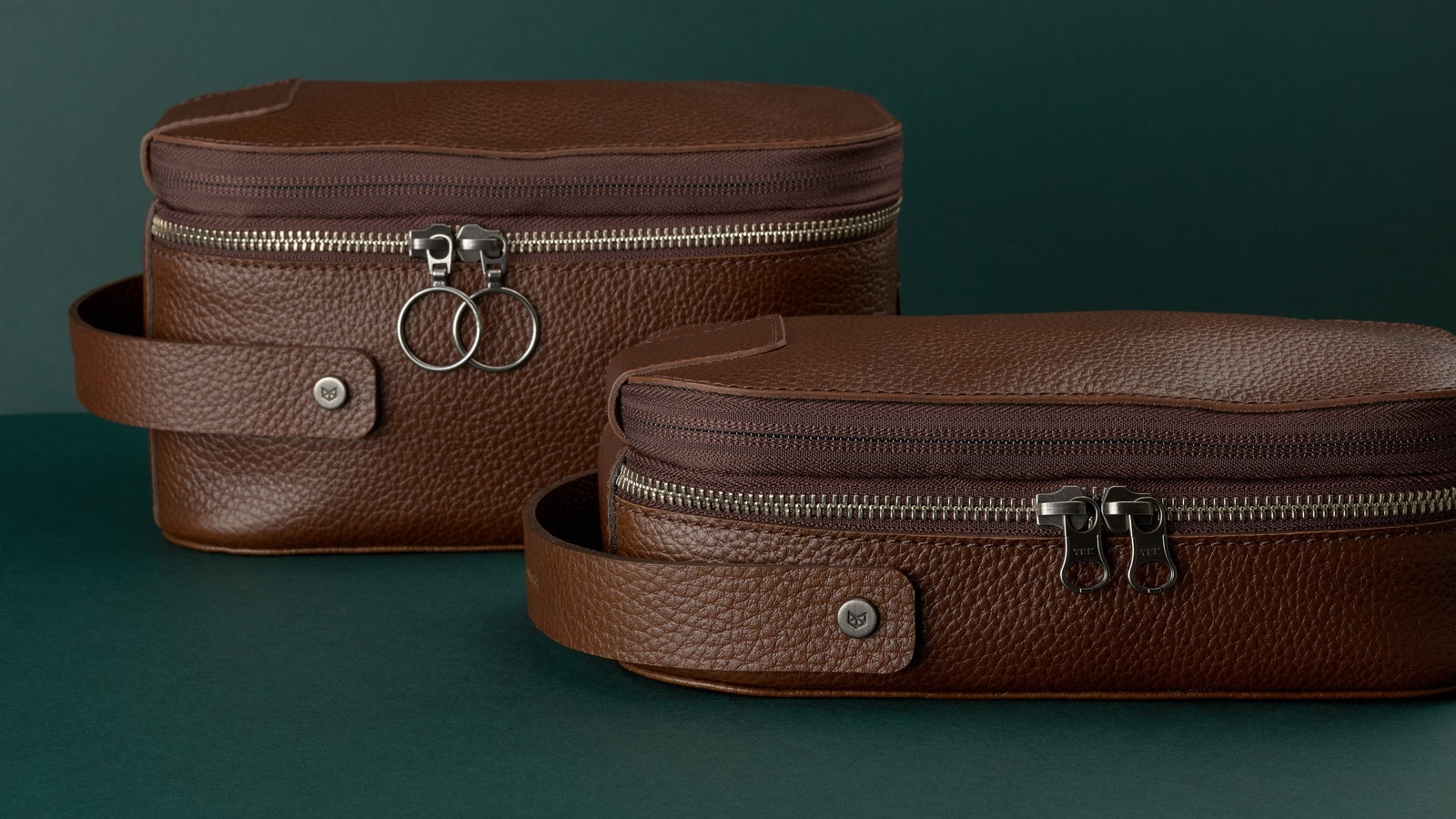 A close-up studio shot featuring two brown toiletry bags, both made of textured, pebble-grain Capra leather.