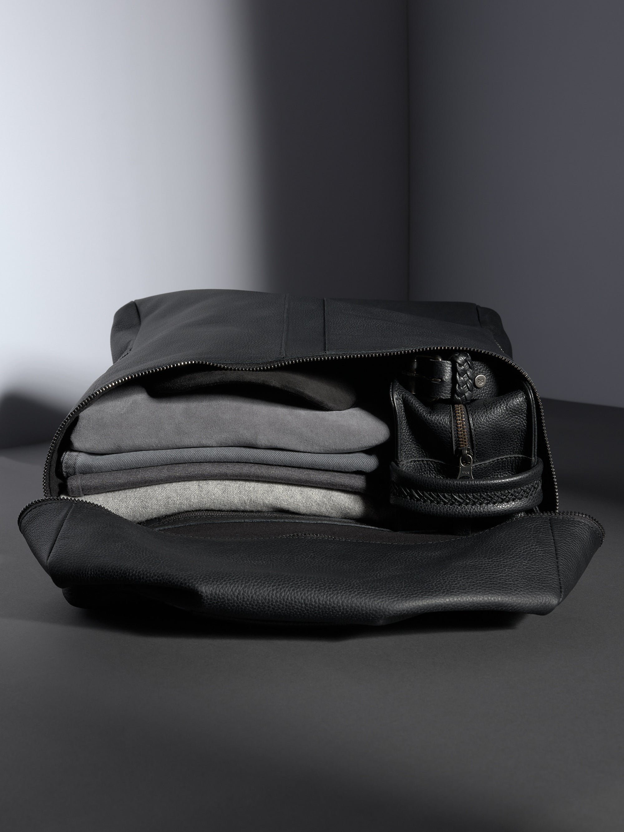 Open Capra Leather Banteng black full-grain leather backpack showing neatly stacked gray clothing and a leather dopp kit.