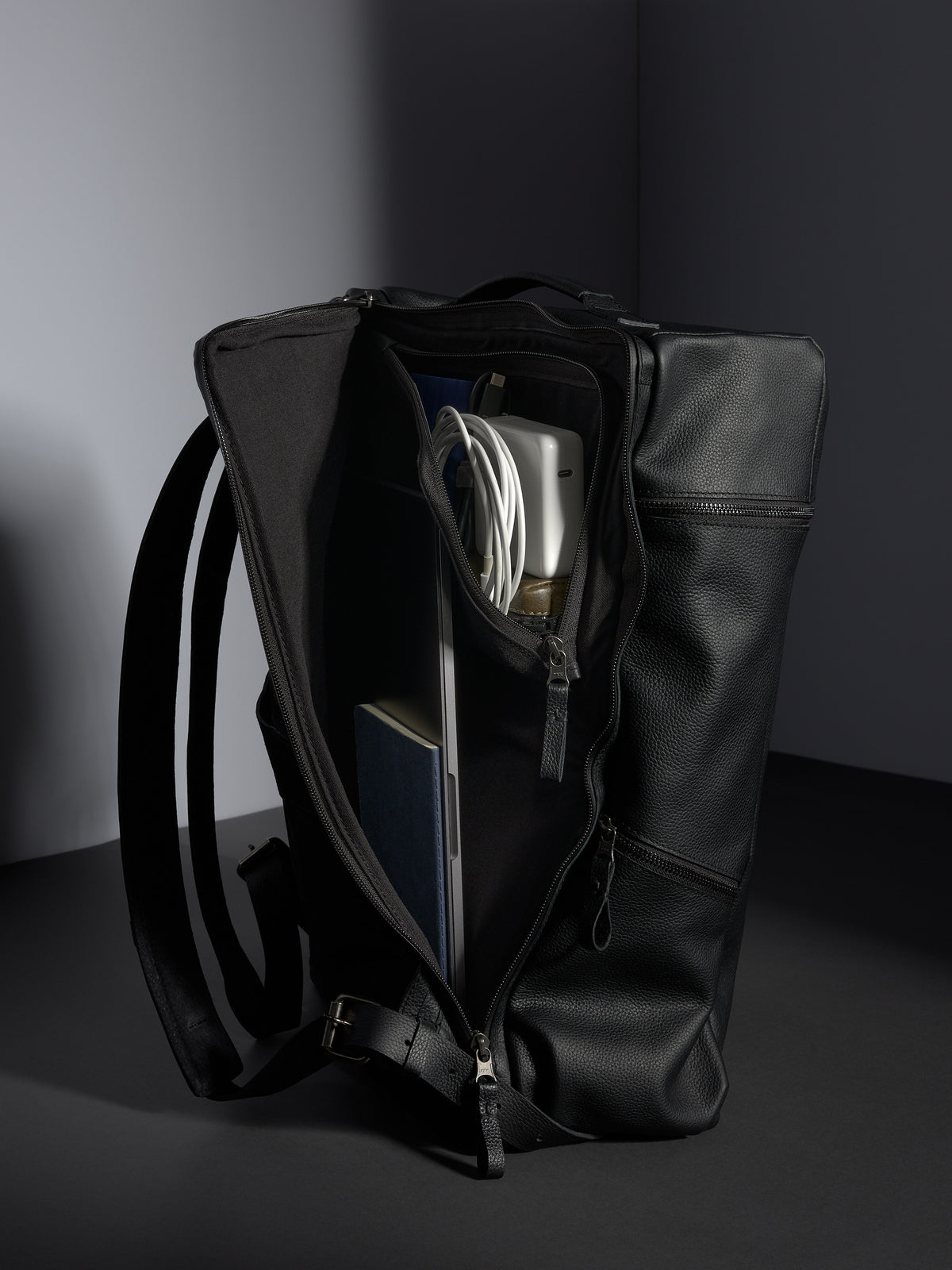 Black Banteng Update leather backpack's main compartment open, showing a laptop, notebook, and power adapter.