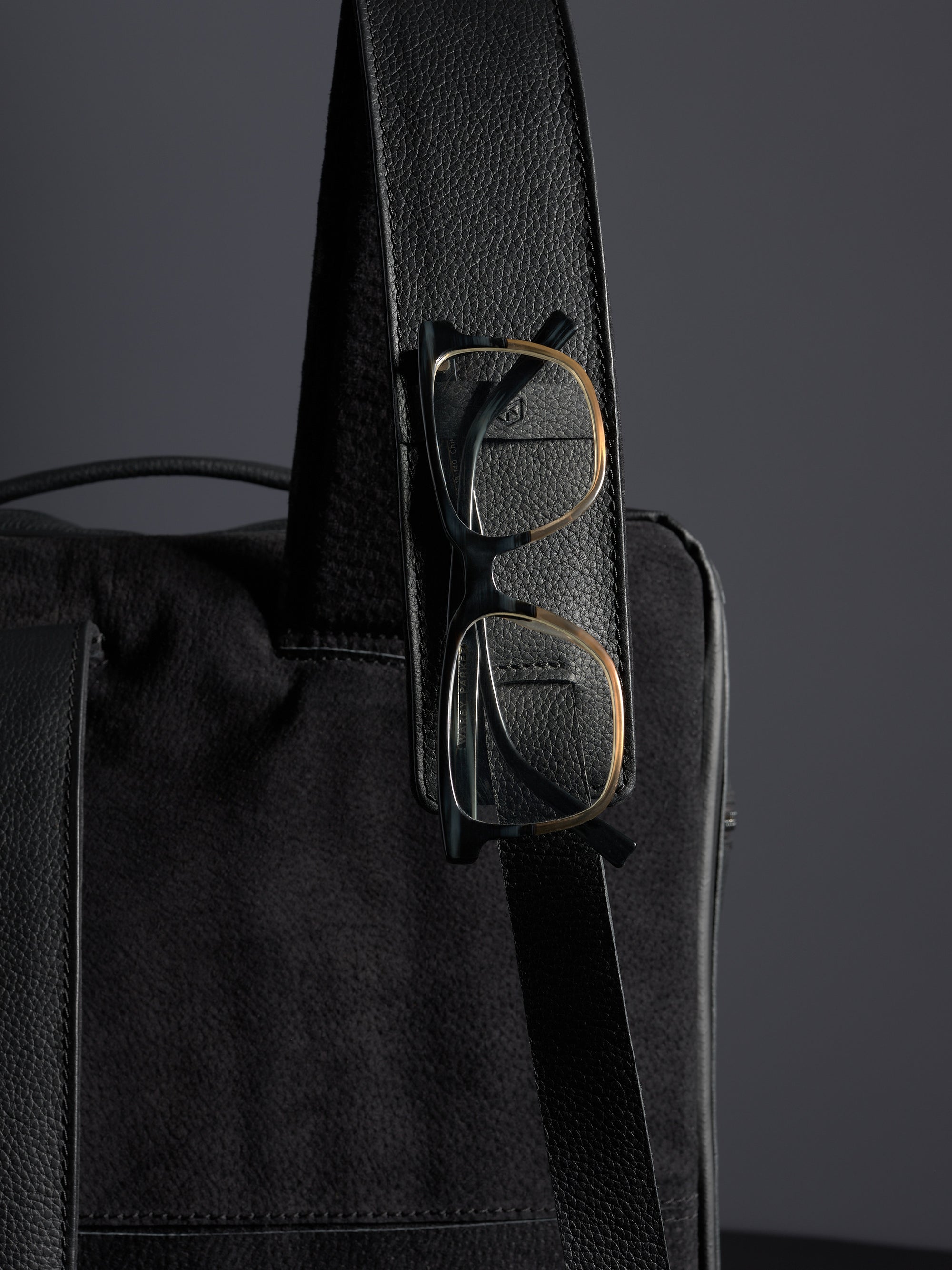 Close-up of Capra Leather black full-grain leather backpack strap with gold and black eyeglasses clipped on.