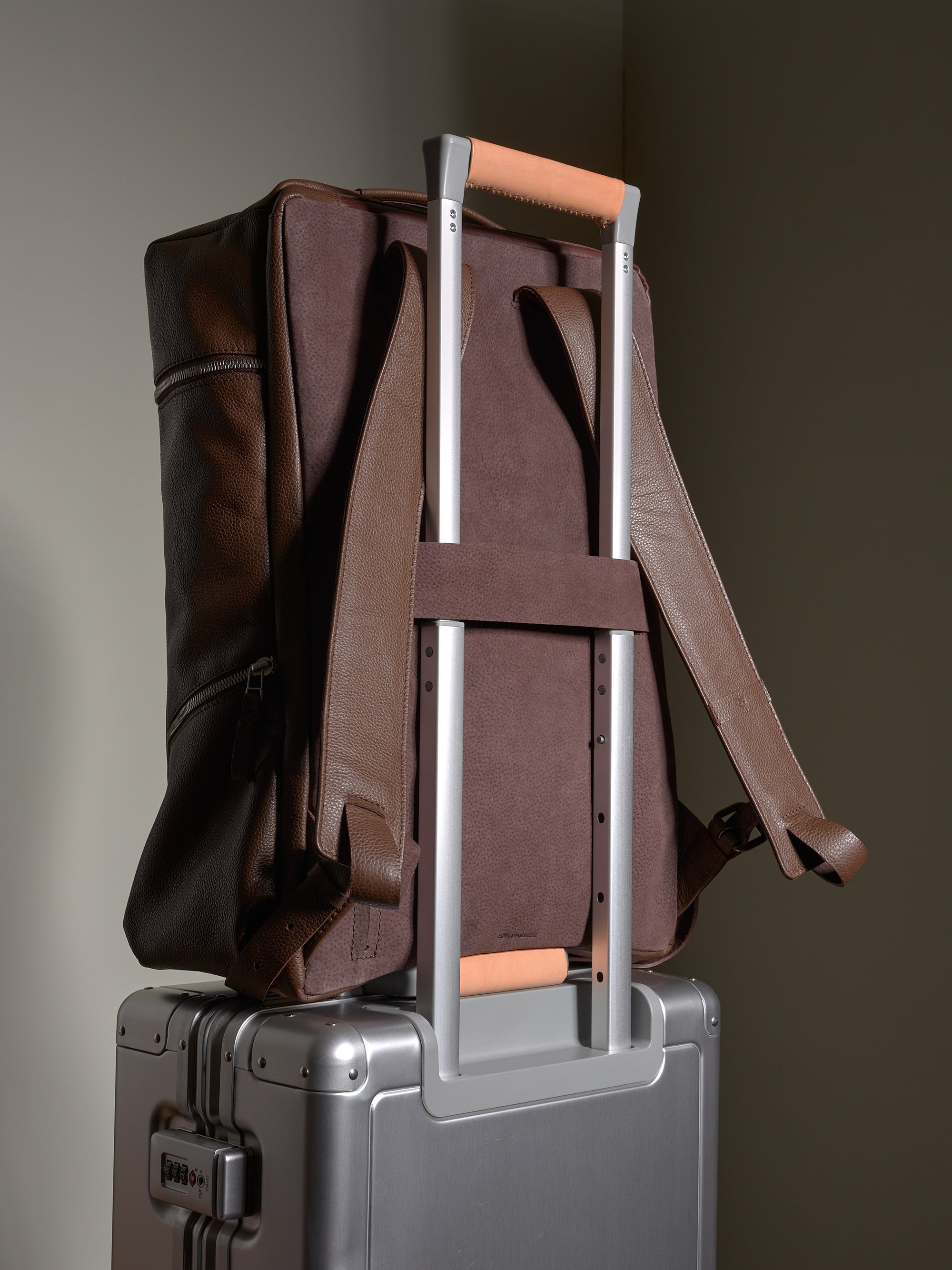 Brown leather backpack with a luggage strap sliding over the handle of a silver rolling suitcase.