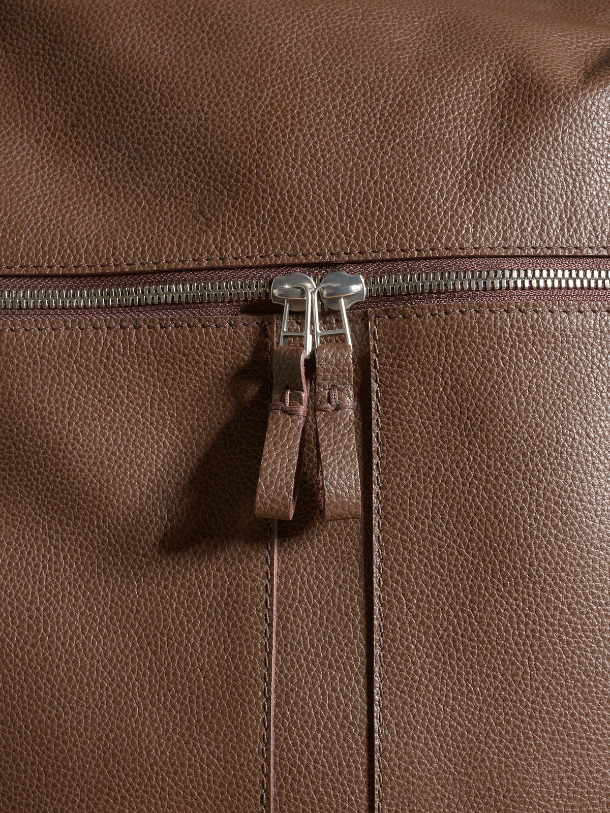 Extreme close-up of the textured brown full-grain leather, stitching, and durable metal zippers.