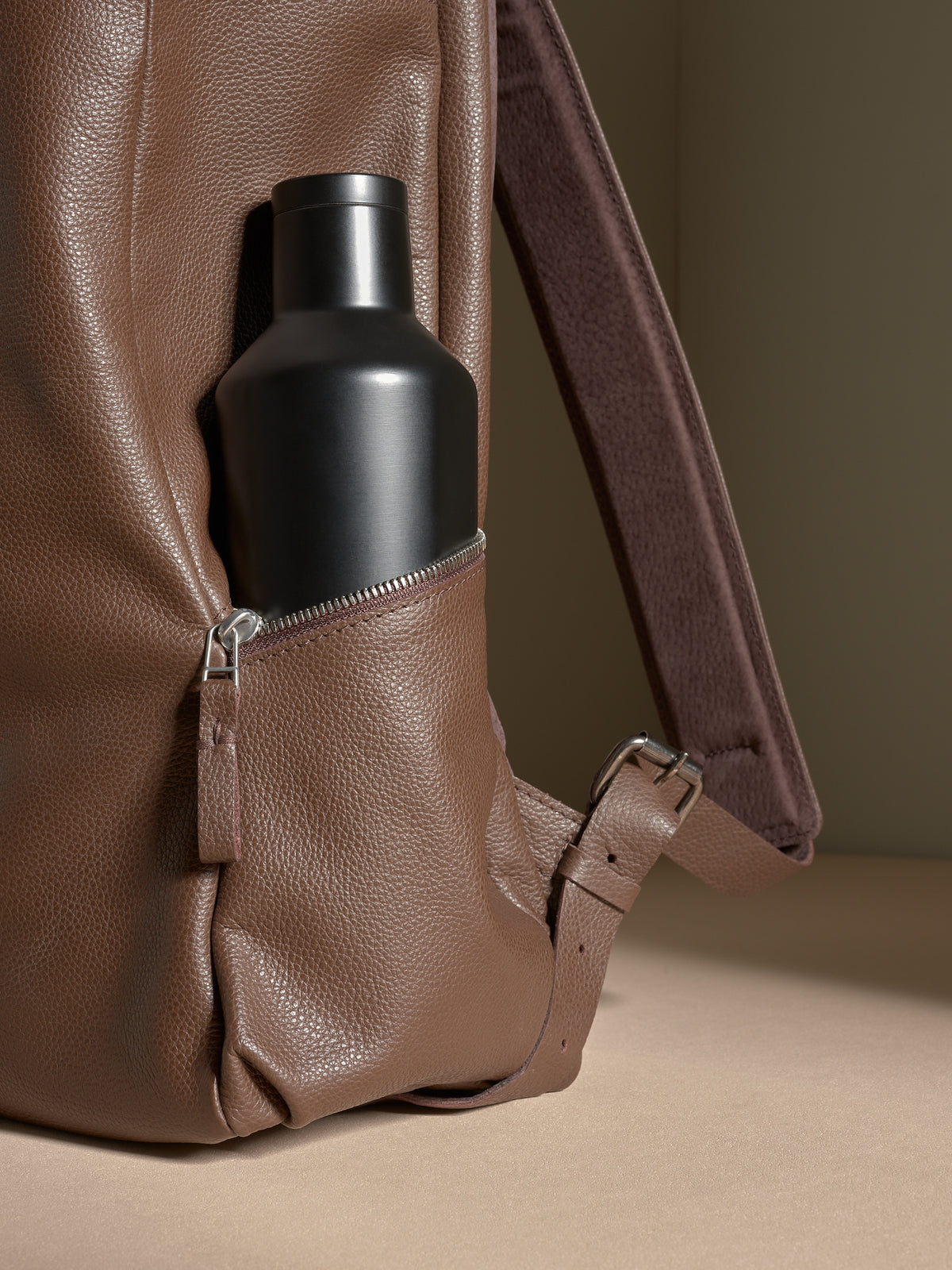 Close-up of a black water bottle secured in the exterior zippered side pocket of a brown leather backpack.