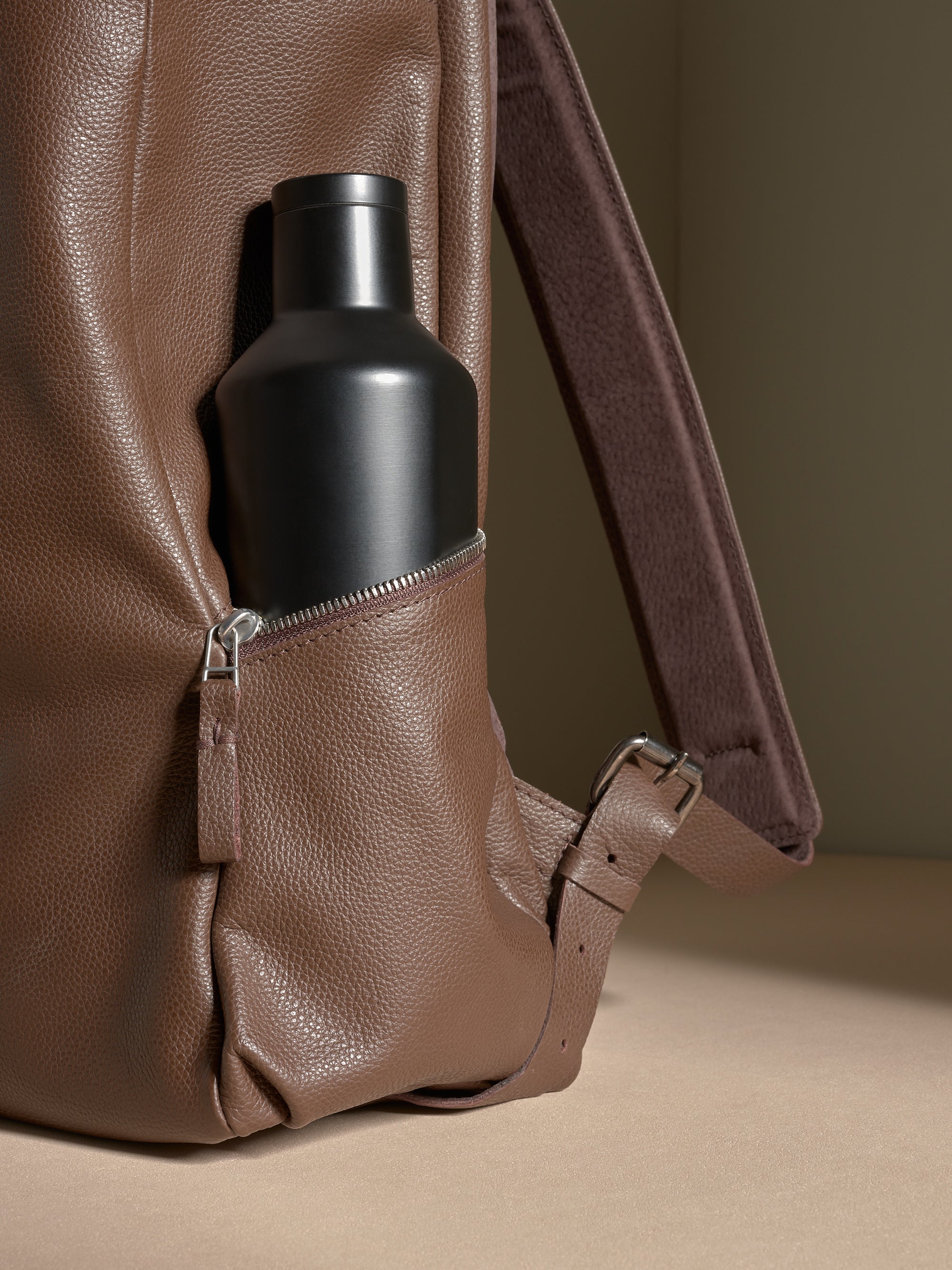 Close-up of a black water bottle secured in the exterior zippered side pocket of a brown leather backpack.