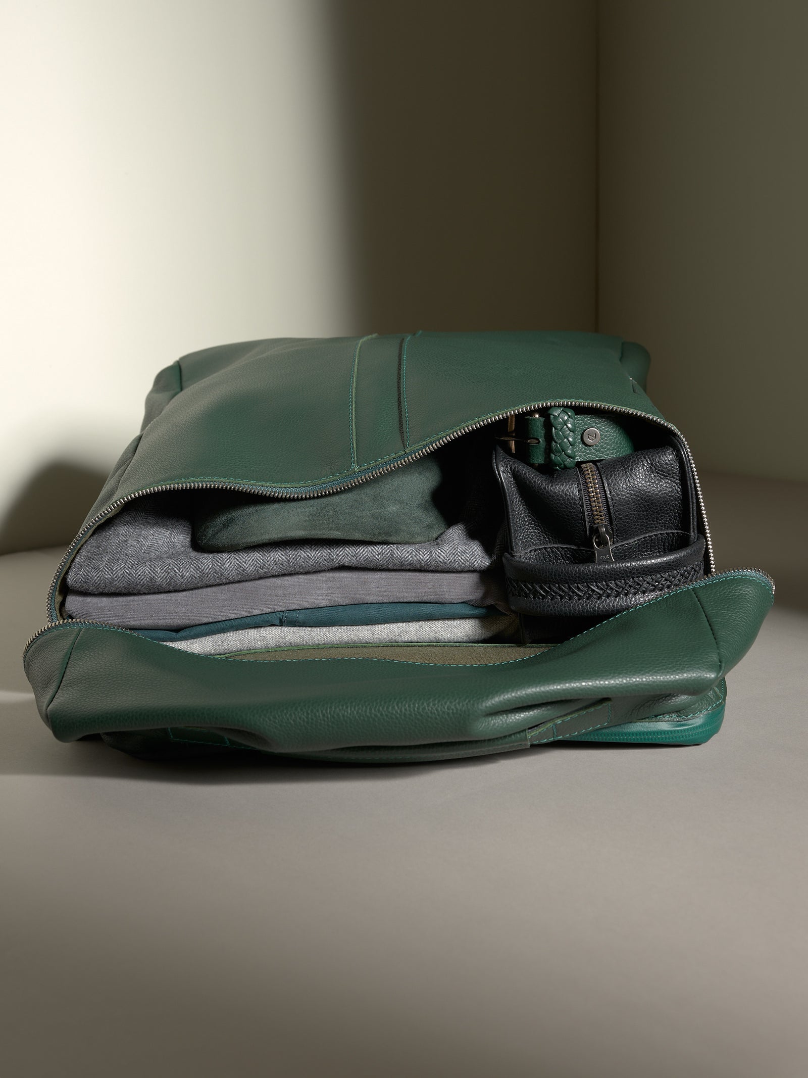 Capra Banteng green leather backpack open, packed for travel with neatly folded clothes and a black leather pouch.