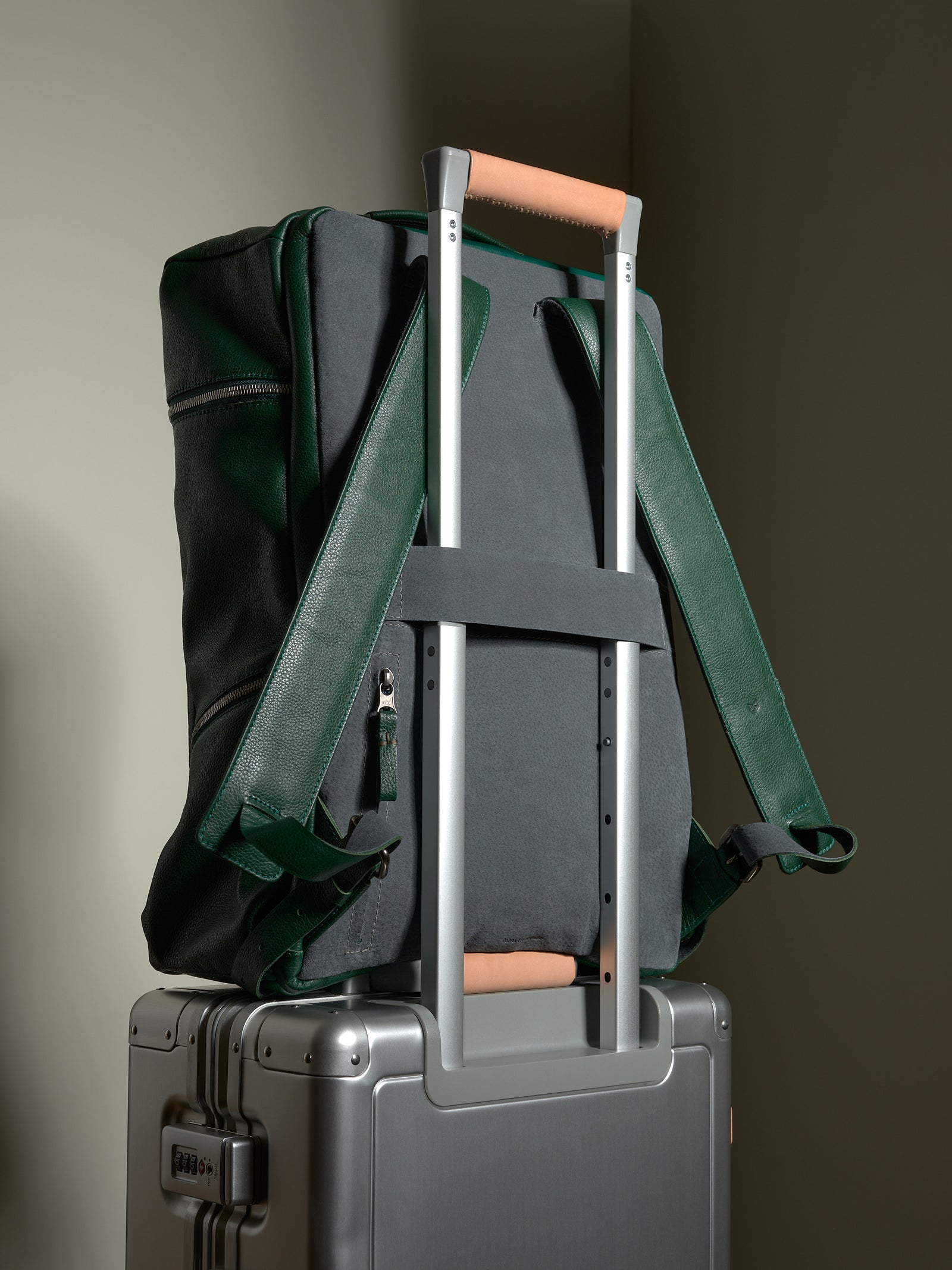 Green leather Capra Banteng backpack resting on a silver suitcase handle, highlighting its travel-ready luggage strap.