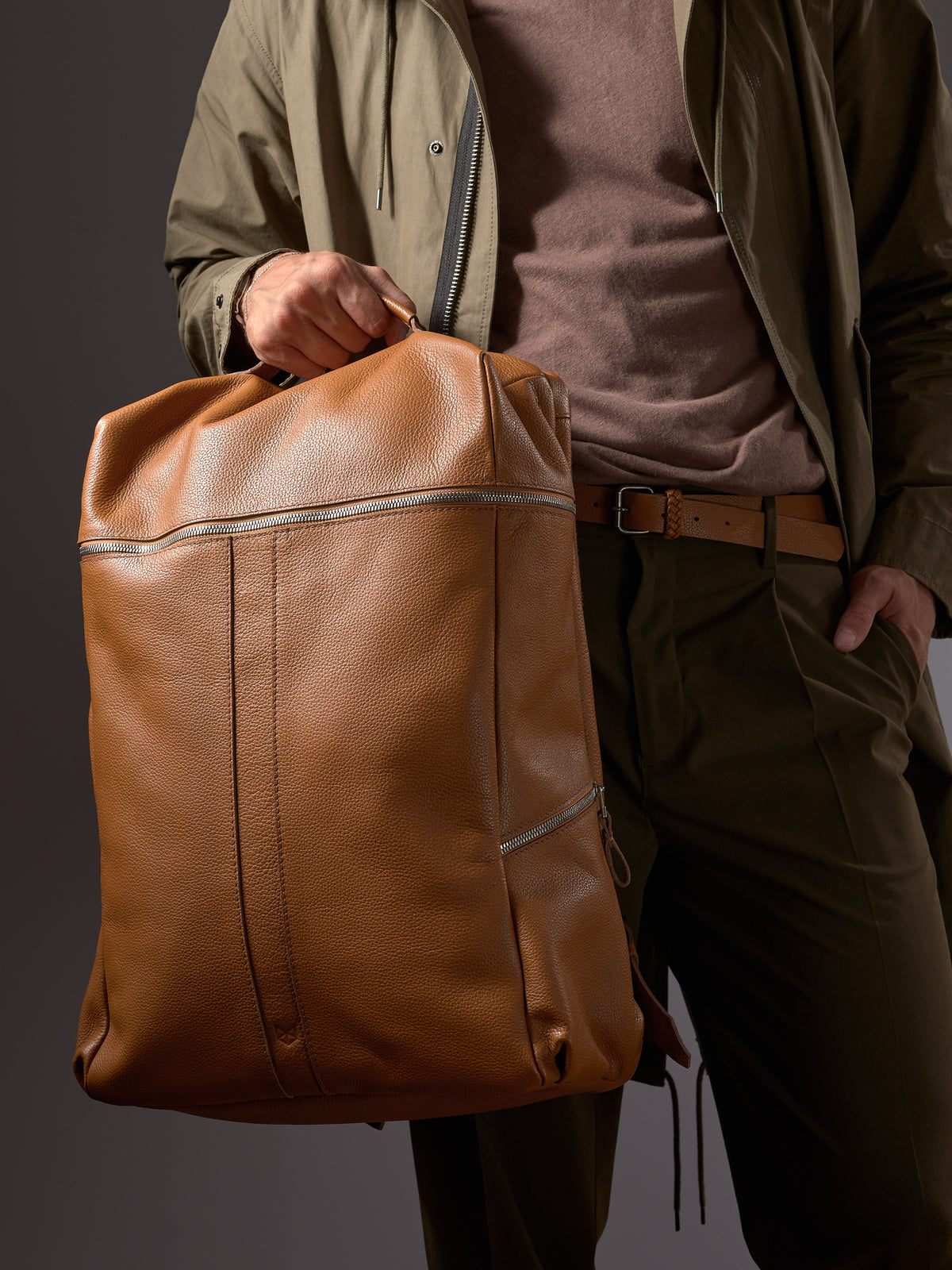 Man holding the Capra Tan Leather Backpack by the handle, showcasing its substantial size and rich color.