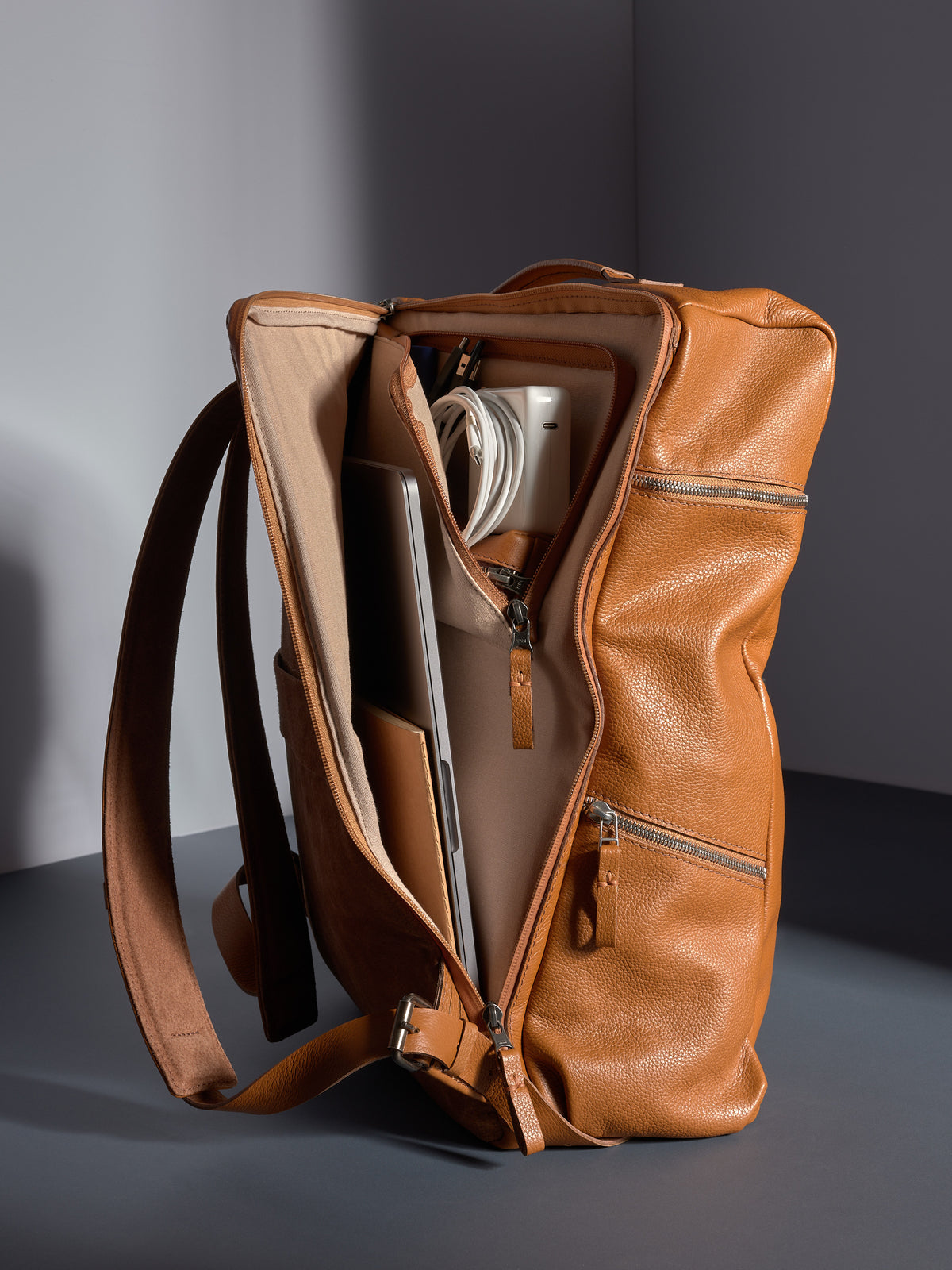 Capra leather backpack opened to show internal storage for a laptop, white charger, and cables.