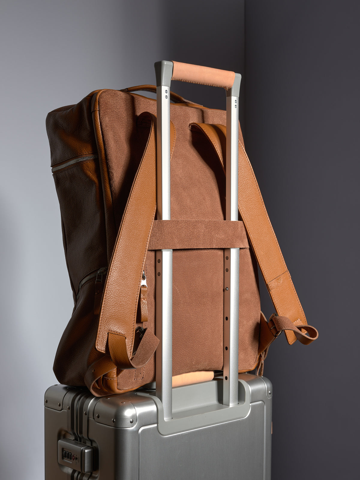 Tan leather backpack secured over a silver rolling suitcase handle using the convenient luggage sleeve.