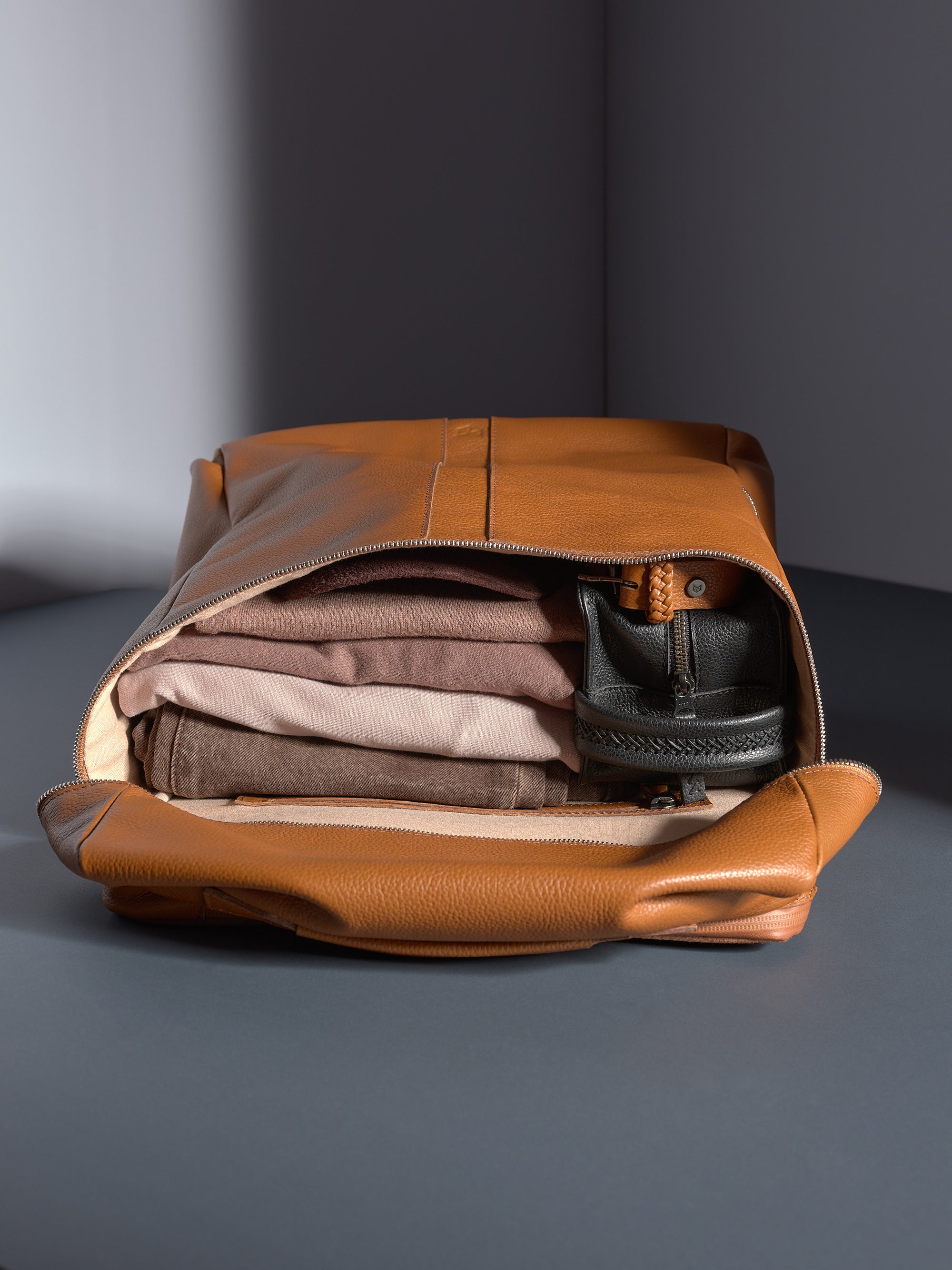 Full-grain tan leather backpack opened, revealing folded brown and beige clothes neatly packed inside.