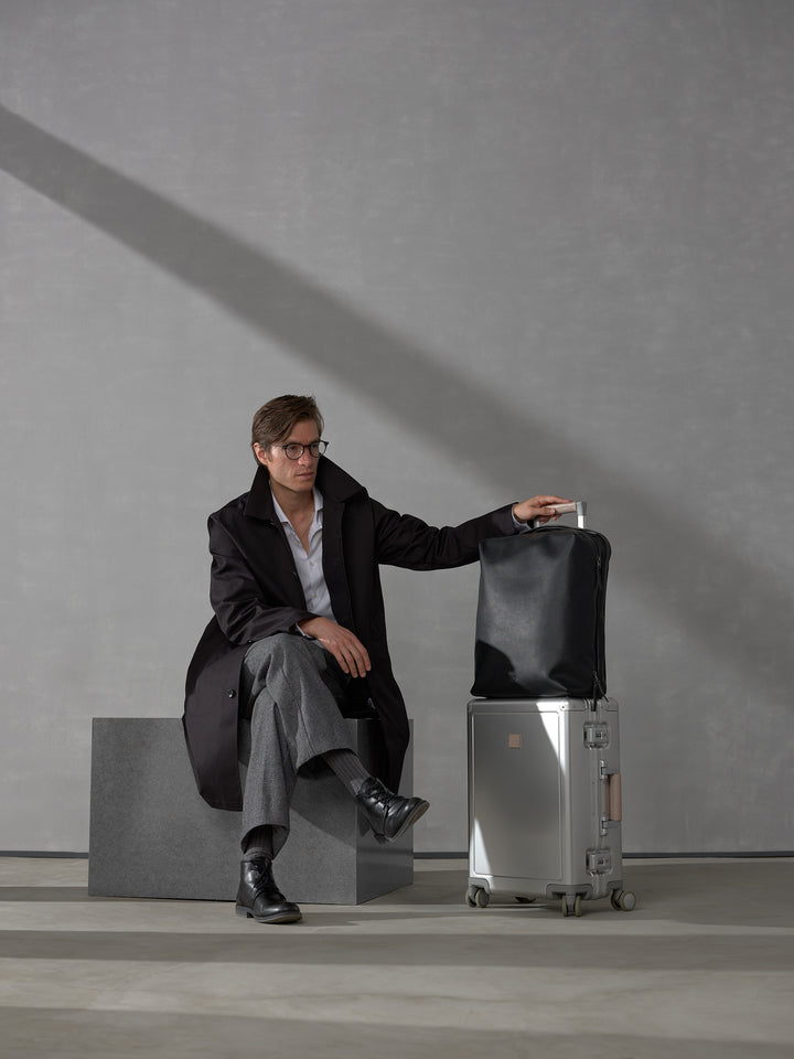 Man in a black trench coat sitting next to a silver suitcase topped with a premium black leather travel backpack.