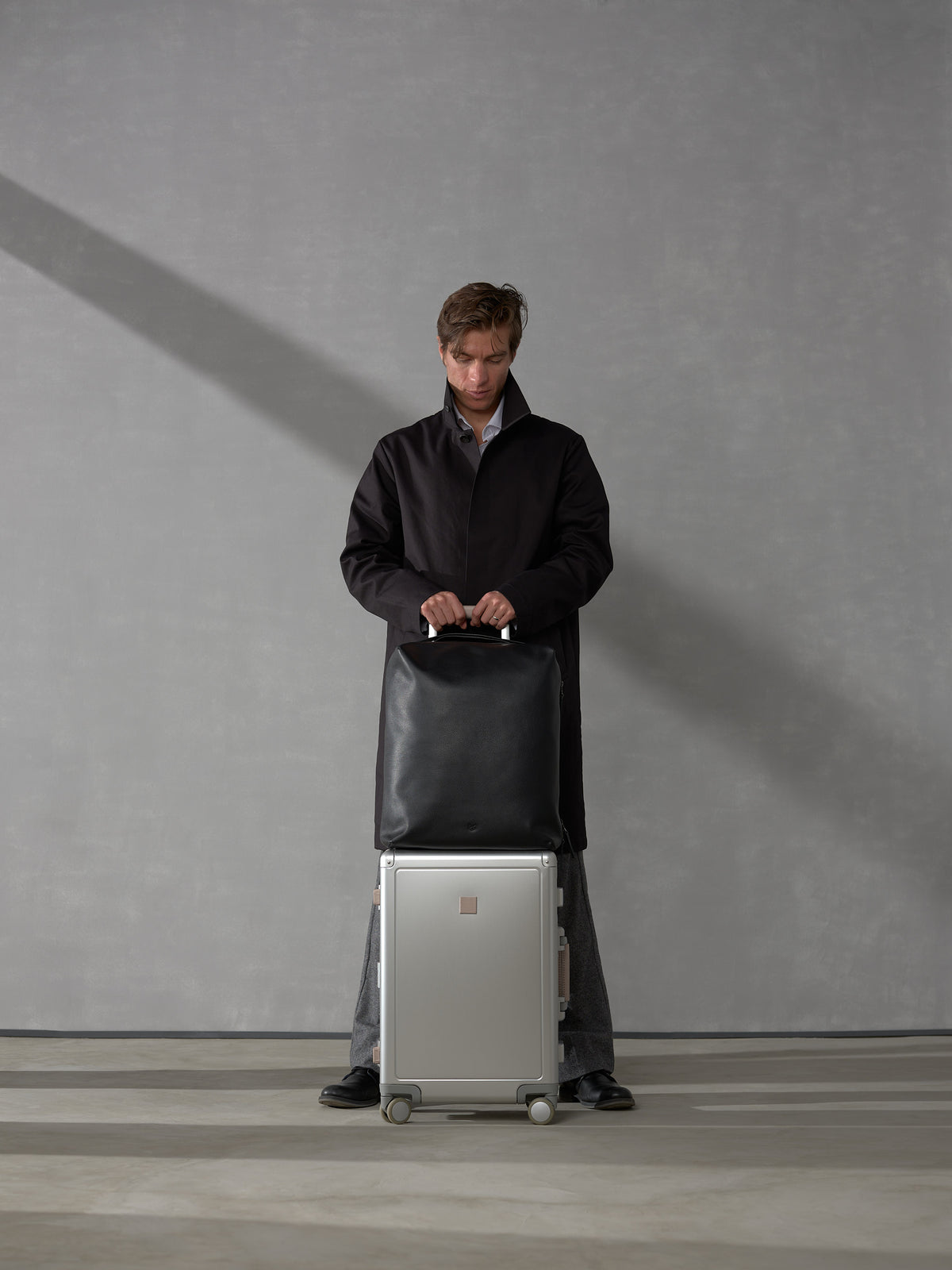 Man in a dark trench coat standing behind a silver rolling suitcase topped with a matching black leather travel bag.