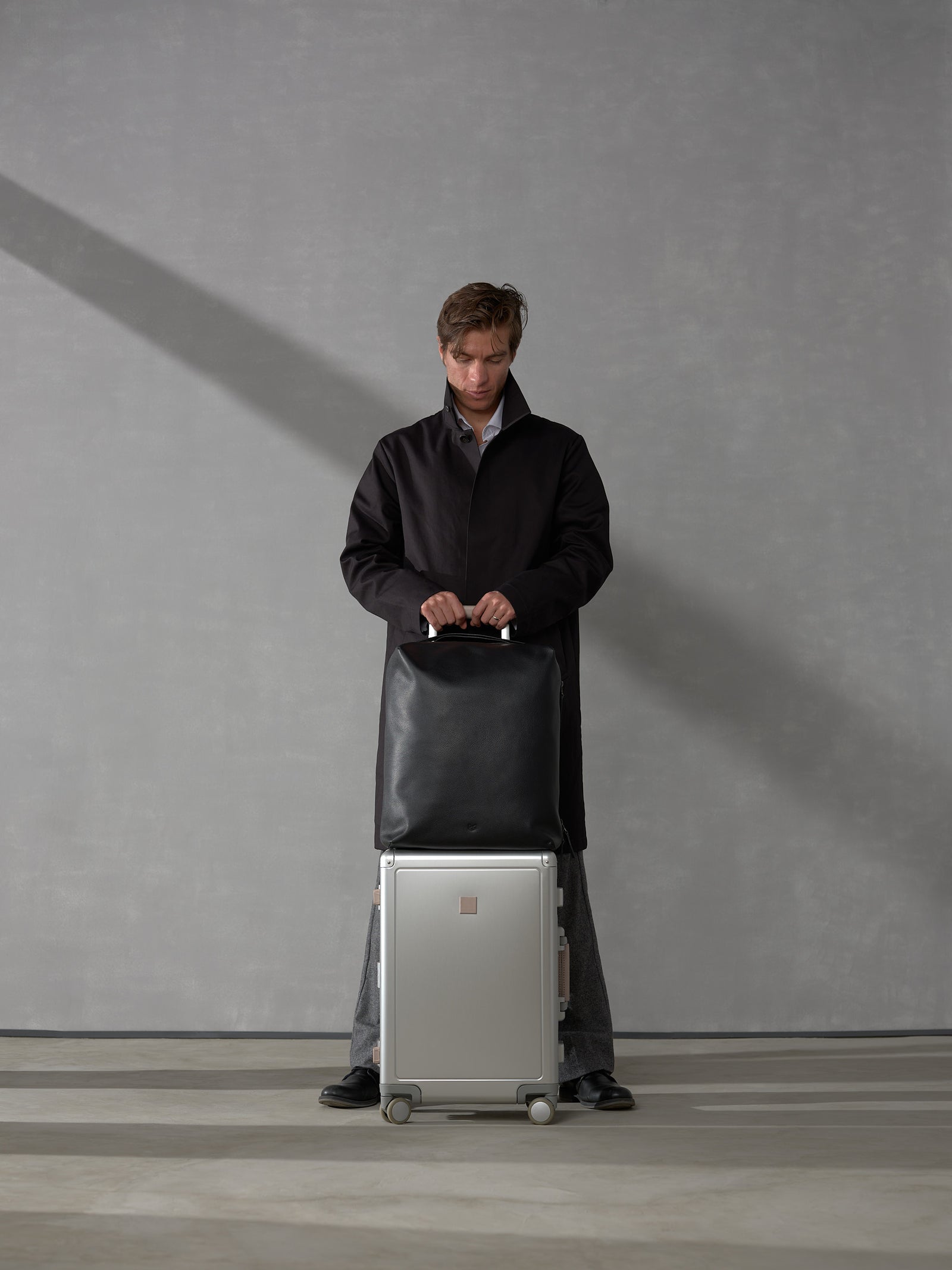 Man in a dark trench coat standing behind a silver rolling suitcase topped with a matching black leather travel bag.