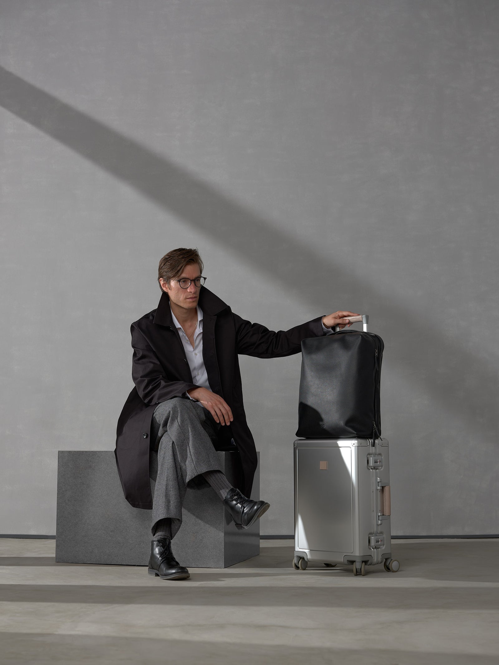 Man in a black trench coat sitting next to a silver suitcase topped with a premium black leather travel backpack.