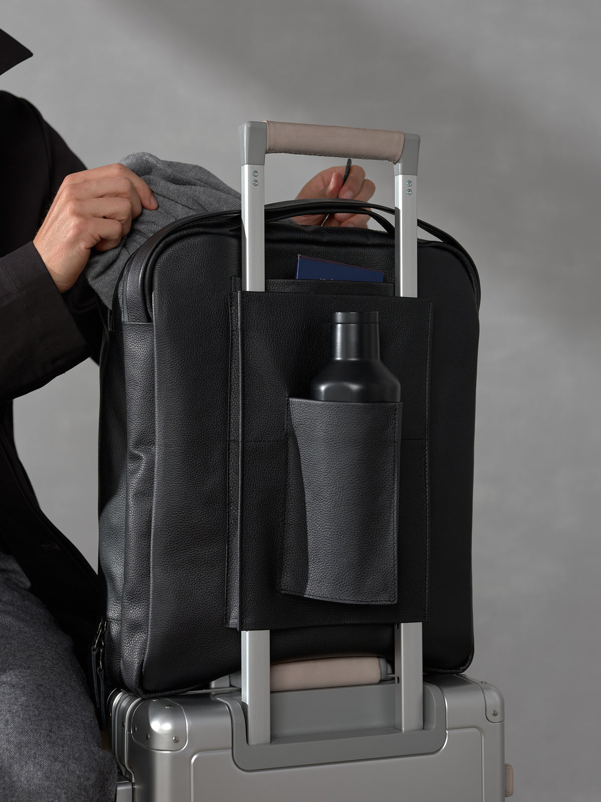 Close-up of a black leather backpack trolley sleeve secured on a suitcase handle, featuring a water bottle pocket.