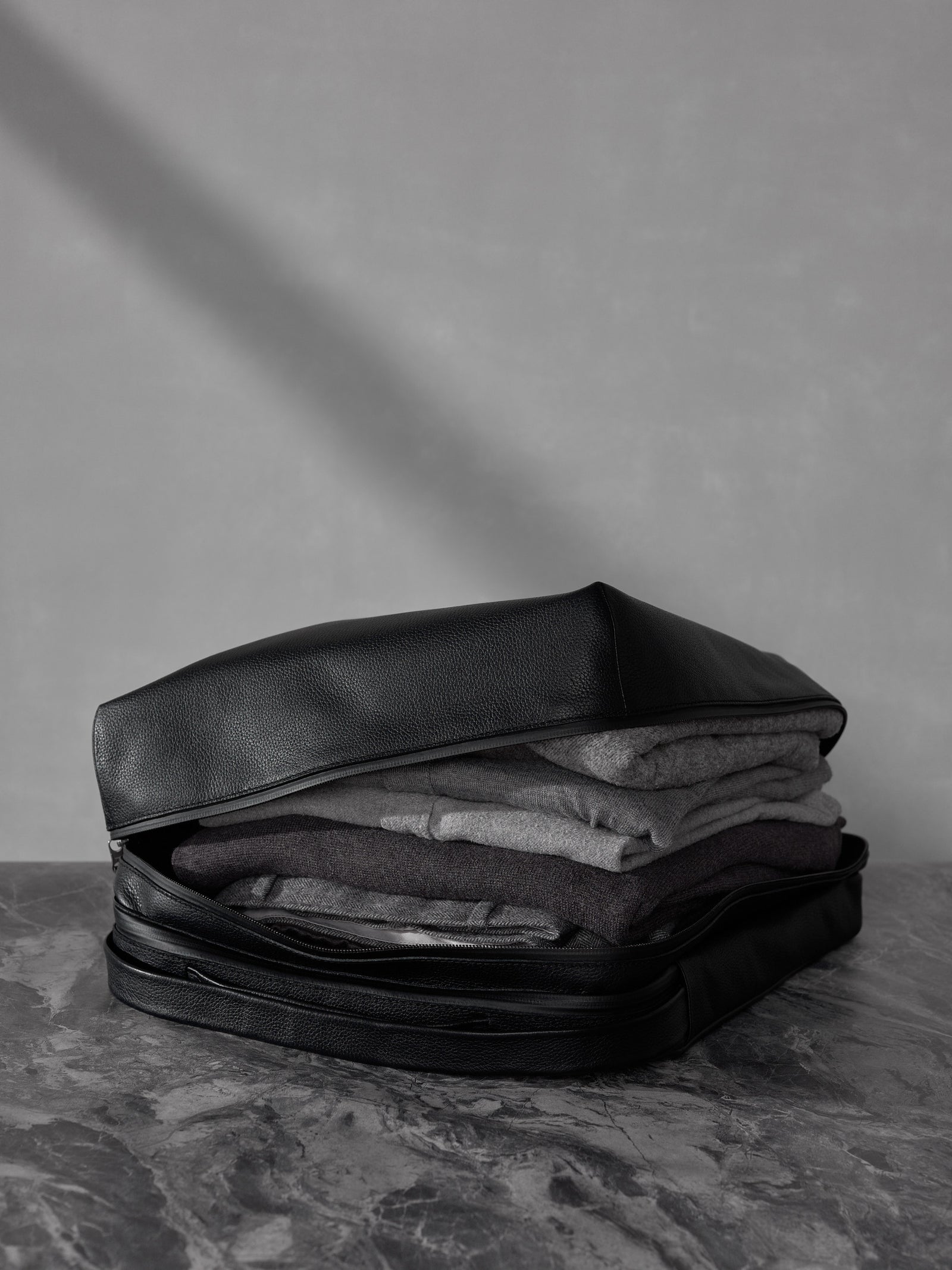 Folded grey sweaters neatly packed inside a black full-grain leather travel bag on a textured stone surface.