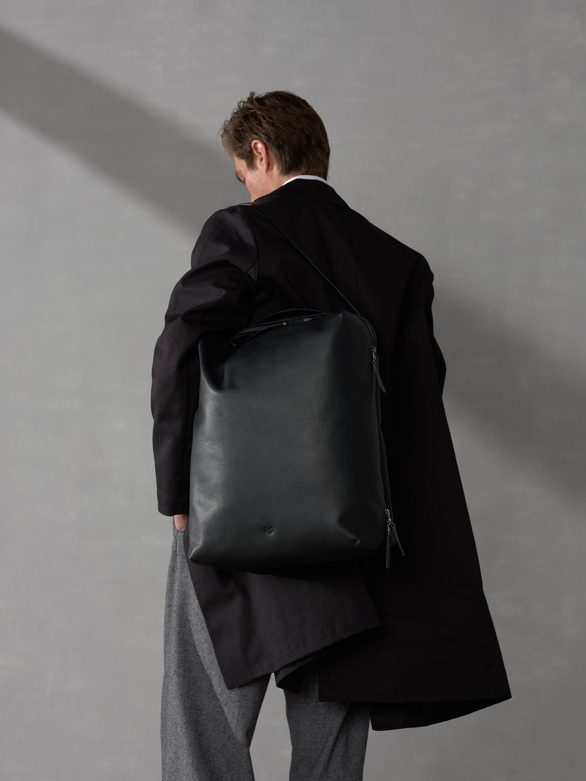 Back view of a man in a dark coat carrying a sleek, minimalist black leather backpack on one shoulder.