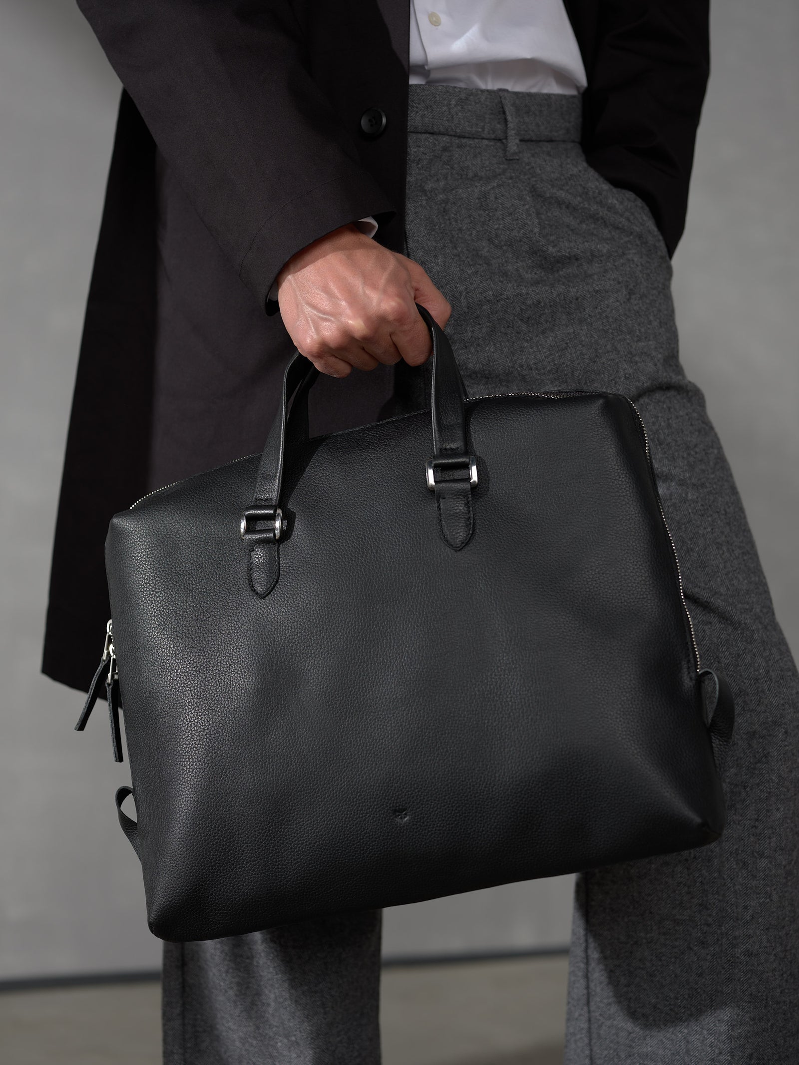 Close-up of a man's hand carrying a sleek black full-grain leather briefcase with premium silver hardware.