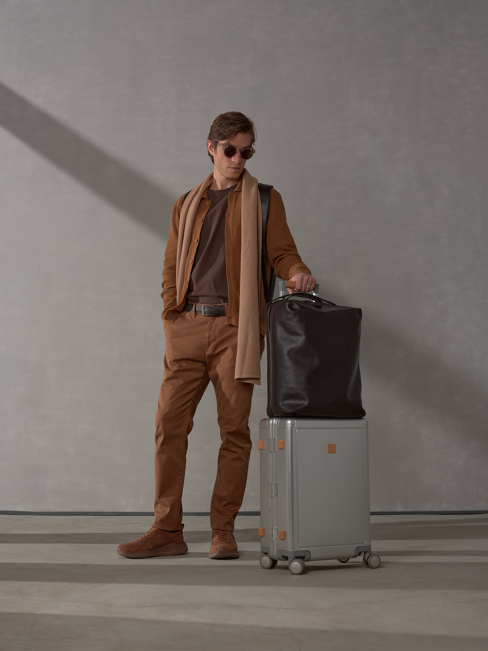 Man in brown stylish clothing standing with Capra Leather backpack attached to a silver rolling suitcase.