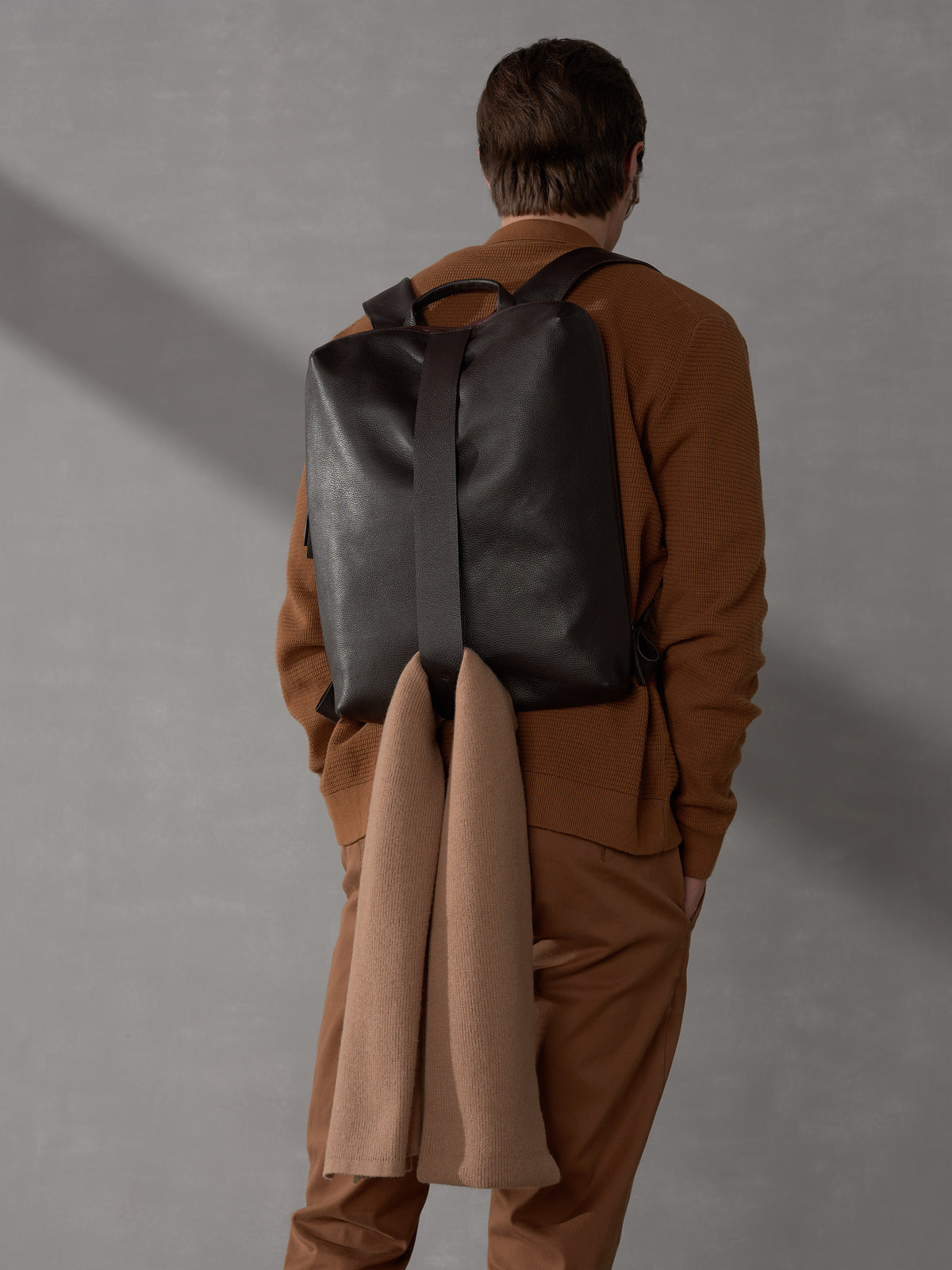 Back view of a man wearing a brown knit shirt and a dark brown luxury leather backpack with a wool scarf.