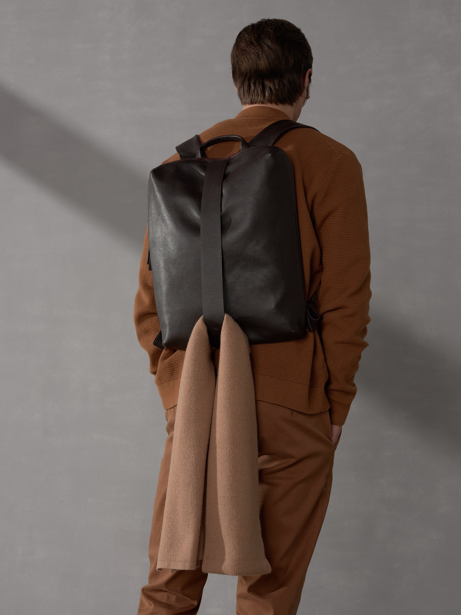 Back view of a man wearing a brown knit shirt and a dark brown luxury leather backpack with a wool scarf.