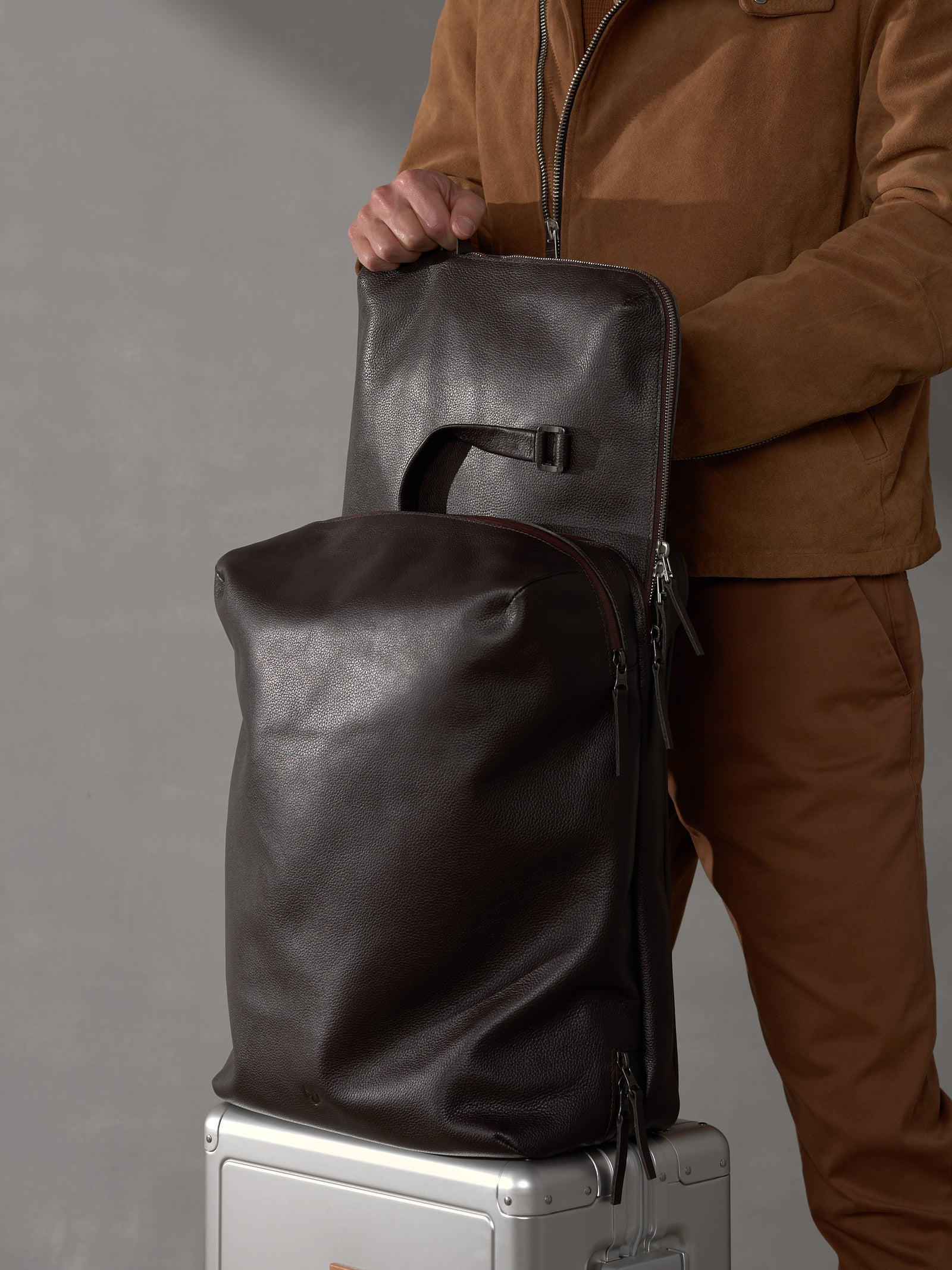 Man detaching a modular component from a large dark brown leather travel backpack system.