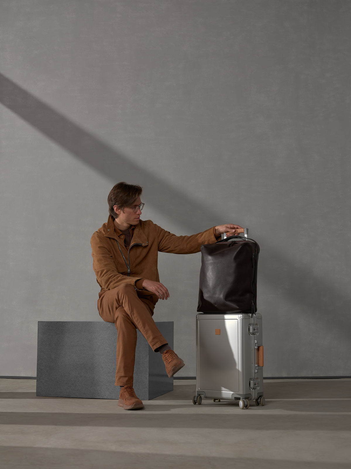 Man in a brown suede jacket sitting down and resting his hand on a silver suitcase with a leather bag on top.