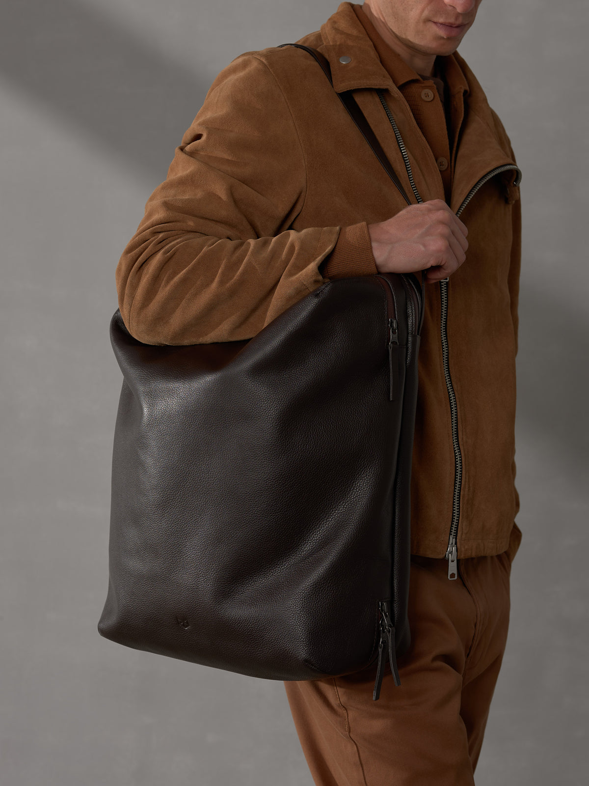 Man wearing a brown suede jacket carrying a large dark brown full-grain leather tote bag over his shoulder.