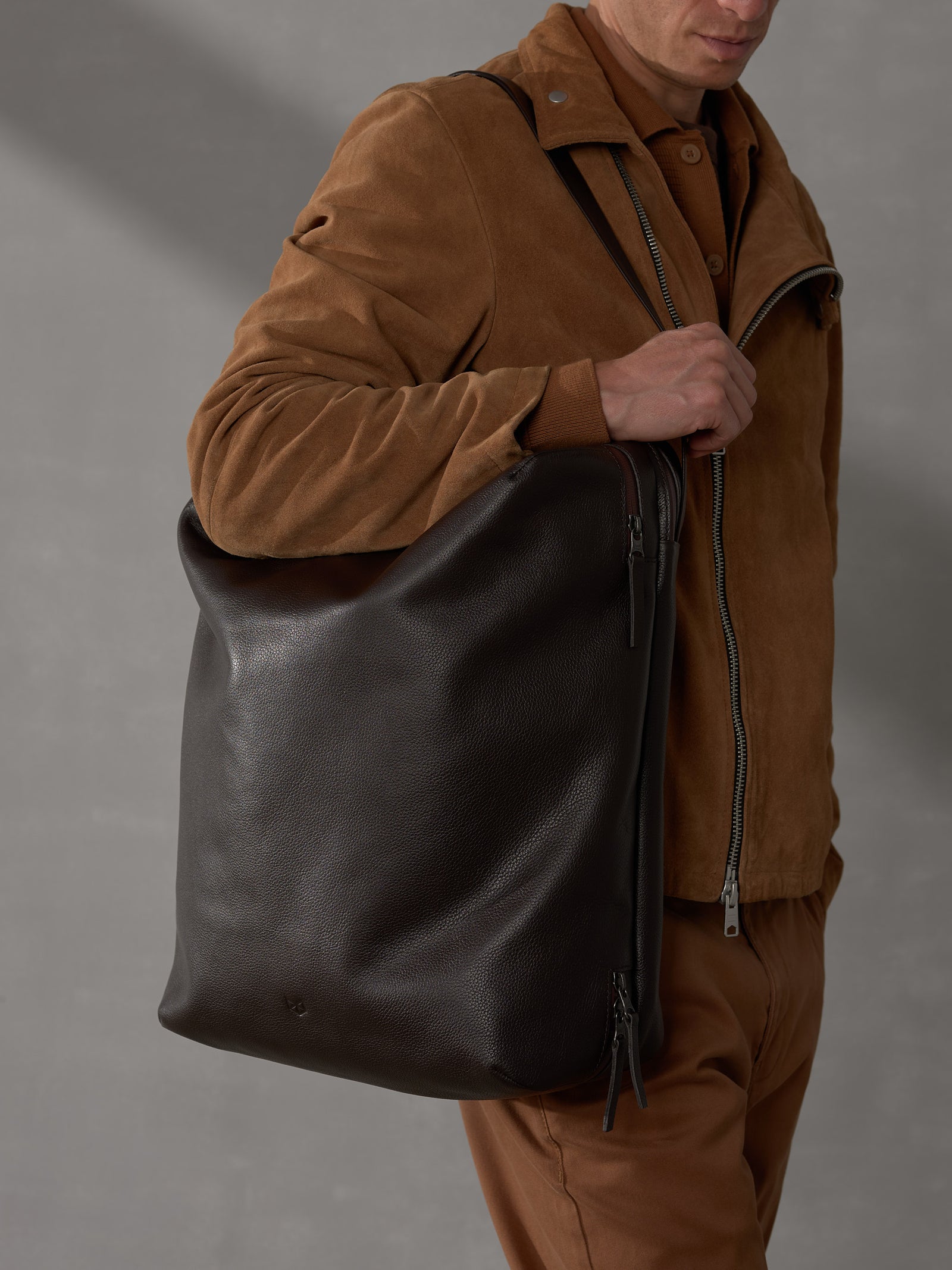 Man wearing a brown suede jacket carrying a large dark brown full-grain leather tote bag over his shoulder.