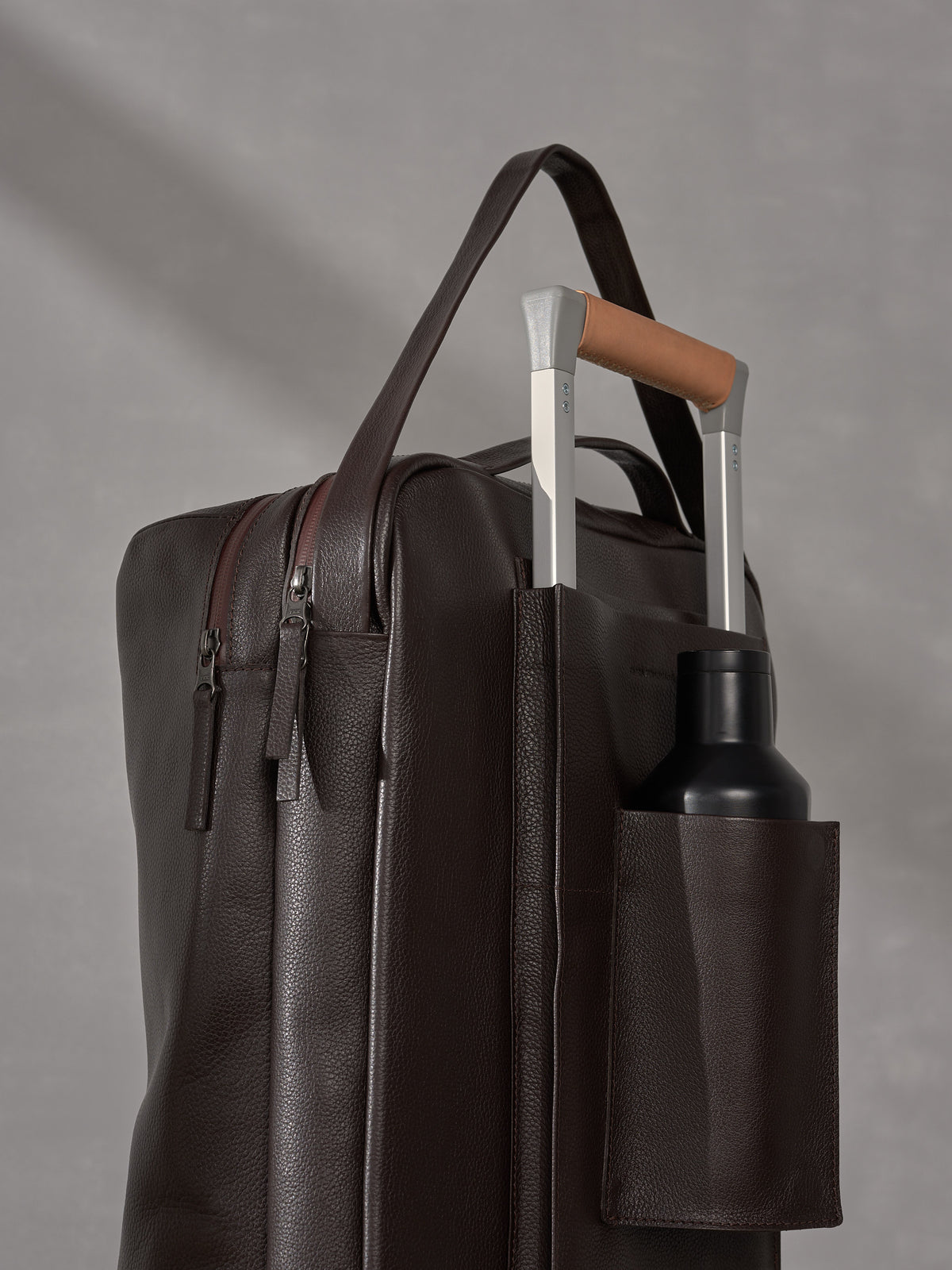 Close-up of a Capra Leather backpack strapped to a luggage handle featuring a built-in water bottle pocket.